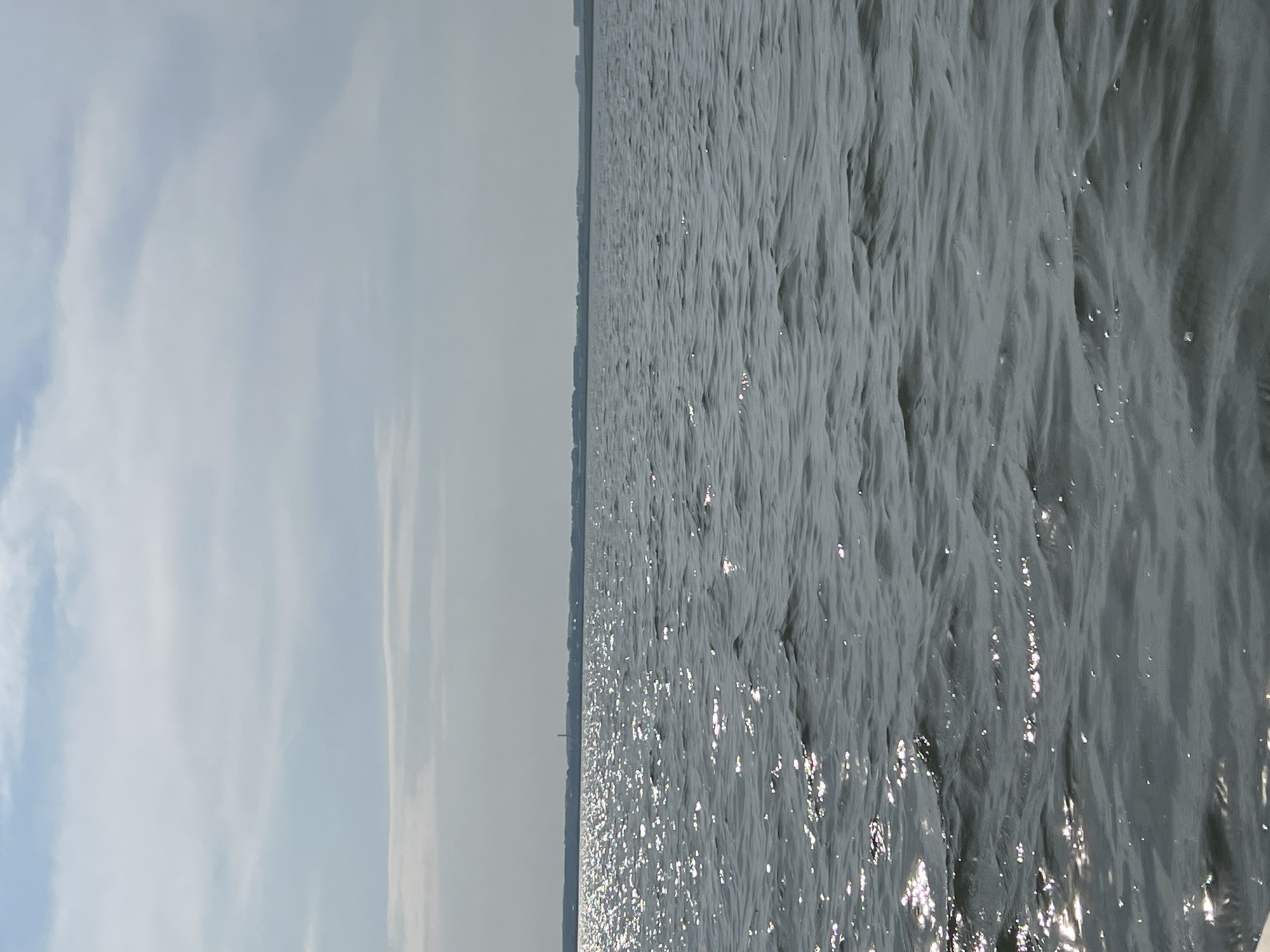 The image depicts a body of water, likely a lake or reservoir, under a partly cloudy sky. The sun is shining, creating bright reflections on the water's surface. In the distance, a tree line is visible, suggesting land along the horizon. The overall impression is one of a tranquil, natural landscape. The provided information about the location in Novopichugovo, Russia, contextualizes the scene as a view of a waterfront area in that specific geographic region. There are no people or specific objects clearly identifiable in the frame beyond the natural elements of water, sky, and distant vegetation.