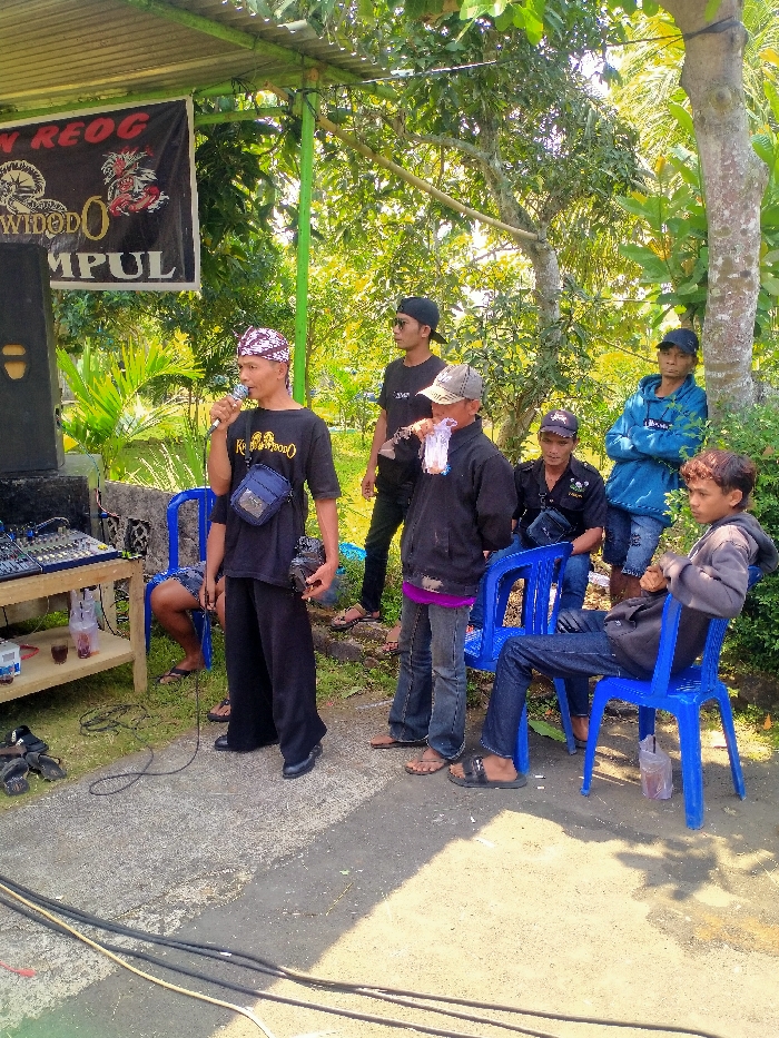 The image depicts an outdoor gathering in Glonggong, Indonesia, under a bright sky with ample natural light. The foreground features a paved ground surface with black and yellow cables running across it. Seven adult males are present in the scene.
On the left, a large black audio speaker stands beside a wooden table holding an audio mixer and several plastic cups containing beverages. Overhead, a black banner with white text, including "WONG REOG" and "WIDODO MPUL," and two stylized dragon-like graphics, is suspended from a green structural pole supporting a corrugated roof.
The central figure, an adult male, stands facing slightly right. He wears a black t-shirt with visible white text, black wide-legged pants, and a traditional brown and white patterned headwrap. He holds a black microphone to his mouth with his right hand and a black camera or similar device in his left, while a dark blue messenger bag is slung across his chest.
To his right, another male, wearing a black jacket, blue jeans, and a grey baseball cap, holds a clear plastic cup with a beverage and straw. Further right and in the background, four additional males are positioned. One is seated on a blue plastic chair, wearing a grey hooded sweatshirt and dark jeans. Another, wearing a blue hoodie and jeans, is seated with his arms crossed. A third is seated on a blue plastic chair wearing a dark shirt and cap. The fourth, wearing a black t-shirt and black baseball cap, stands behind the group. Lush green trees and foliage form the background, indicating a verdant, semi-rural setting.