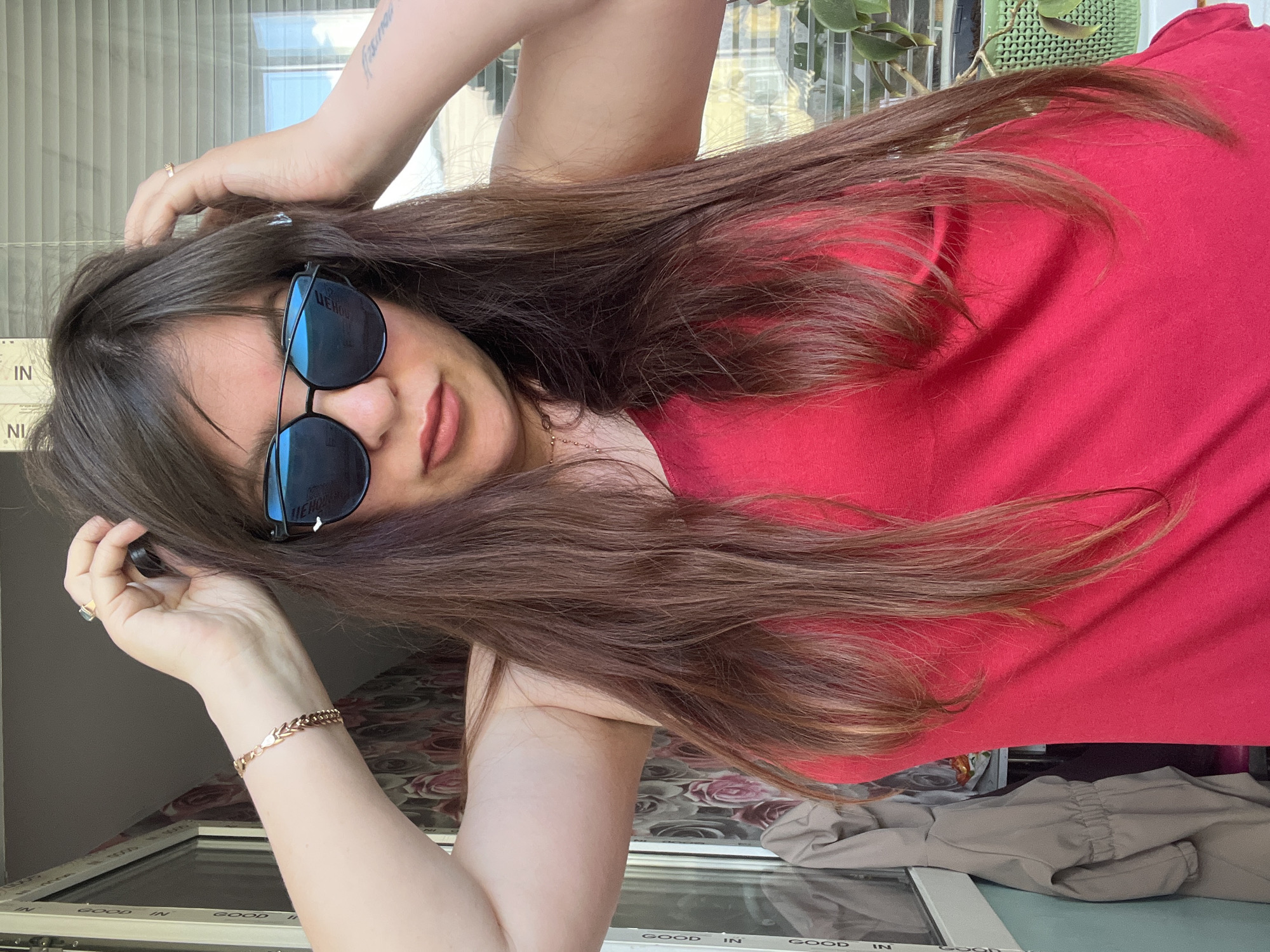 In Noviy, Russia, a woman with long brown hair and sunglasses is posing with her hands on her head in front of a window. She is wearing a red tank top. A bracelet is visible on her left wrist, and a ring is on her right index finger. The background shows a window with blinds and a plant.