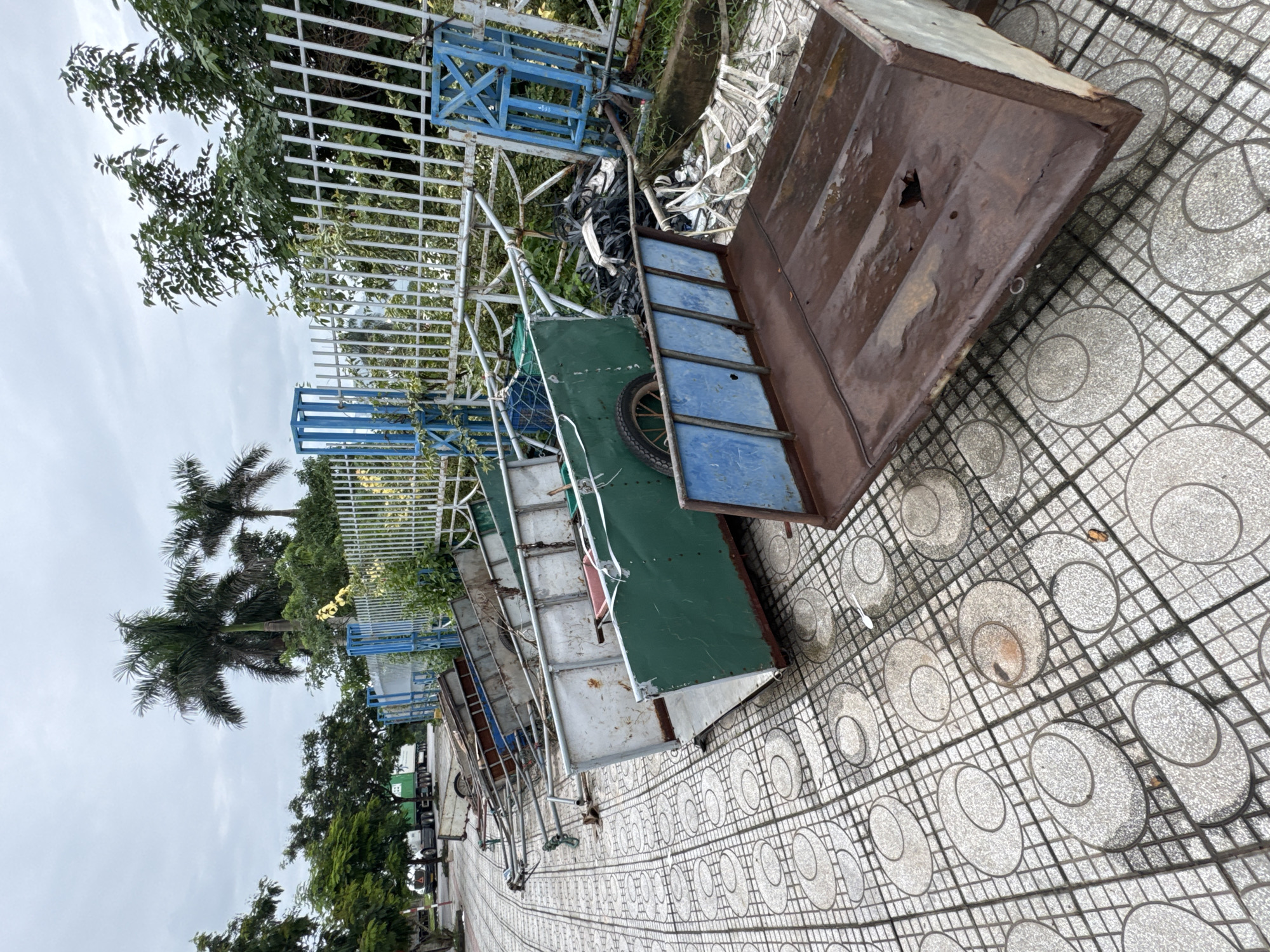 The image depicts a scene in Lao Bảo Commune, Vietnam, likely an outdoor area with a tiled walkway and a fence. Several metal carts, appearing to be abandoned or in disrepair, are scattered on the ground. Some are green and white, while others are made of rusted metal. A white metal fence with blue accents runs along the right side of the image, partially obscured by overgrown vegetation. Tall palm trees are visible in the background, suggesting a tropical or subtropical climate. The sky is overcast, and the ground appears wet in places, indicating recent rain. There are no people visible in the frame. The overall impression is one of neglect and abandonment.