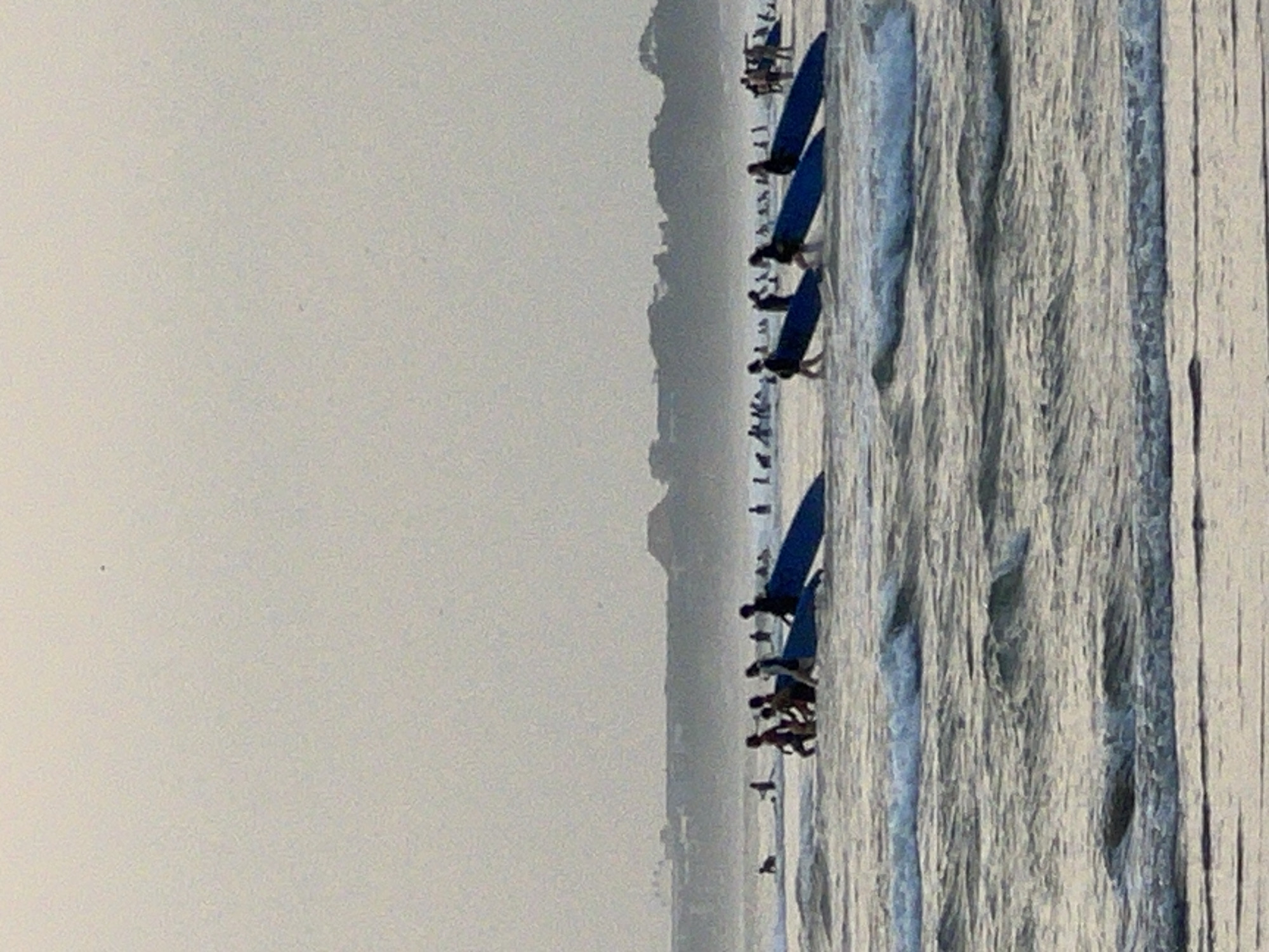 This is a medium-shot, horizontal, grainy, outdoor photograph taken during daylight, likely at dusk due to the hazy, muted light. The scene depicts a group of people, many carrying surfboards, wading in the shallow waters of Kuta Beach, Bali, Indonesia. The ocean is choppy with small waves breaking. In the foreground, the water is a muted grey-blue, with foamy white caps. Further back, a line of about 8-10 individuals, appearing to be a mix of adults and possibly younger people, are walking in the water, some carrying blue surfboards. The surfboards are positioned upright and angled slightly towards the viewer. Behind them, other individuals and surfboards are visible, scattered further out in the water. The distant background shows a hazy, dark green treeline and buildings, likely the Kuta coastline, under a pale grey, overcast sky. The overall impression is of a surfing lesson or a group heading out to surf.