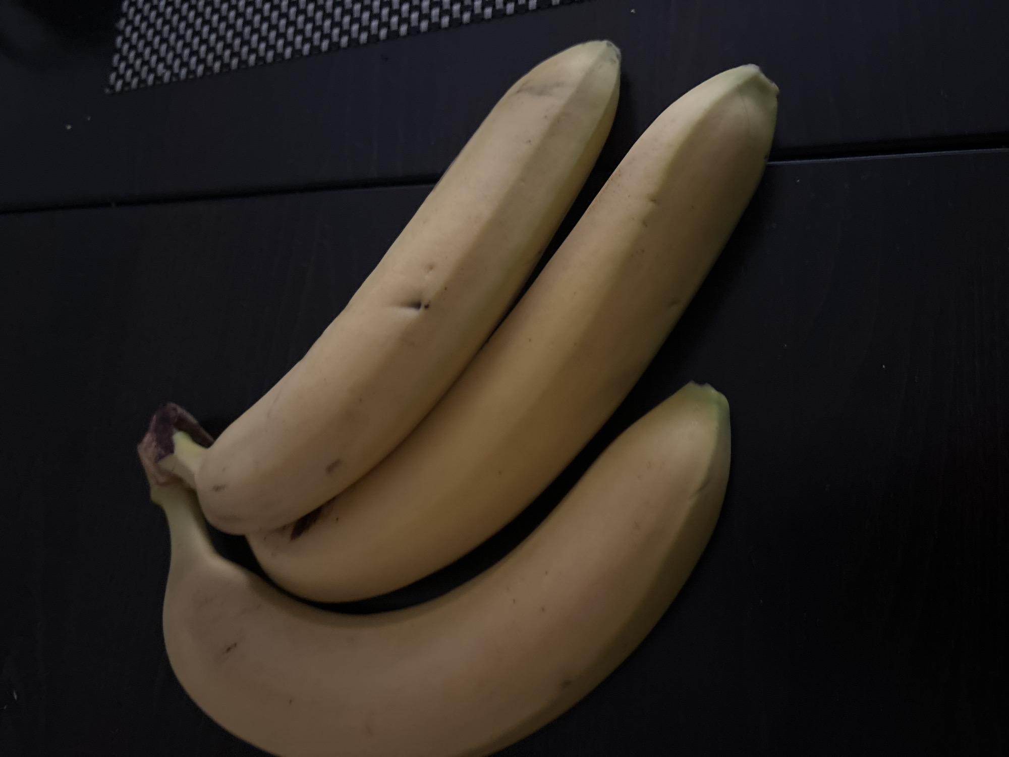 Three bananas are placed on a dark surface. The bananas are ripe and have a yellow color with some brown spots. The surface appears to be a table or countertop. The location, Budva, Montenegro, is not evident from the image.
