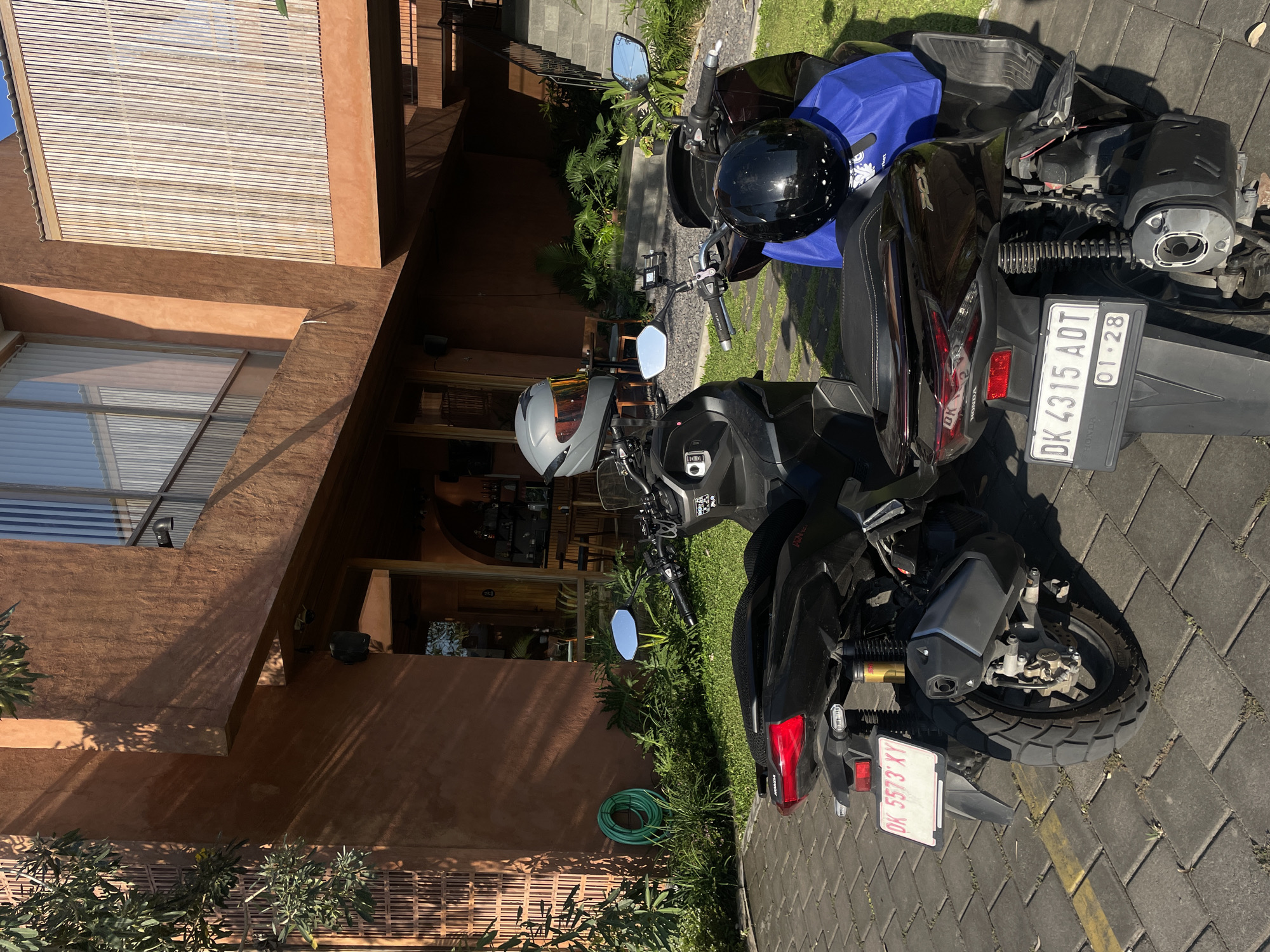 Two dark-colored scooters are parked on a paved patio area in front of a building with a warm, terracotta-colored exterior. The scooters are positioned side-by-side, with the rear ends facing the camera. The scooter on the left has a silver helmet with an orange visor on its handlebars and a license plate that reads "DK 5573 XY". The scooter on the right has a black helmet and a blue bag on its seat, and its license plate reads "DK 4315 ADT" with "01.28" below it.
Behind the scooters, a lush green garden area with various plants and trees is visible, including a coiled green garden hose on the wall. The building features a large window with vertical blinds and a wooden slatted facade on an upper level. To the right of the building, an outdoor staircase leads up to a higher floor. The scene is illuminated by bright sunlight, casting shadows on the pavement and building. The image does not contain any people or indicate any specific activities. The location is not specified as Kediri, Indonesia.