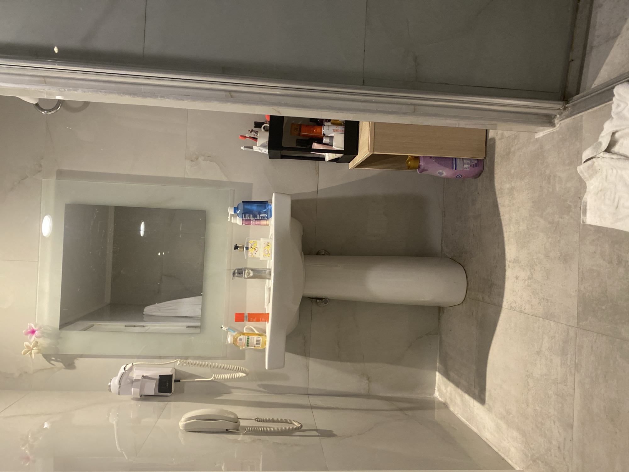 The image shows a bathroom scene in Aksu, Turkey. A white pedestal sink with a chrome faucet is the central focus, positioned in front of a large rectangular mirror. To the left of the sink, a white wall-mounted hairdryer and a telephone are visible. On the countertop of the sink, there are several items including bottles of toiletries, a toothbrush holder with toothbrushes, and a bar of soap. To the right of the sink, a black organizer unit filled with various beauty products and toiletries sits on a small wooden cabinet. A large bottle of purple liquid, possibly shampoo or body wash, is on the floor next to the cabinet. The floor is tiled in a light gray color. The scene is illuminated by overhead lighting.