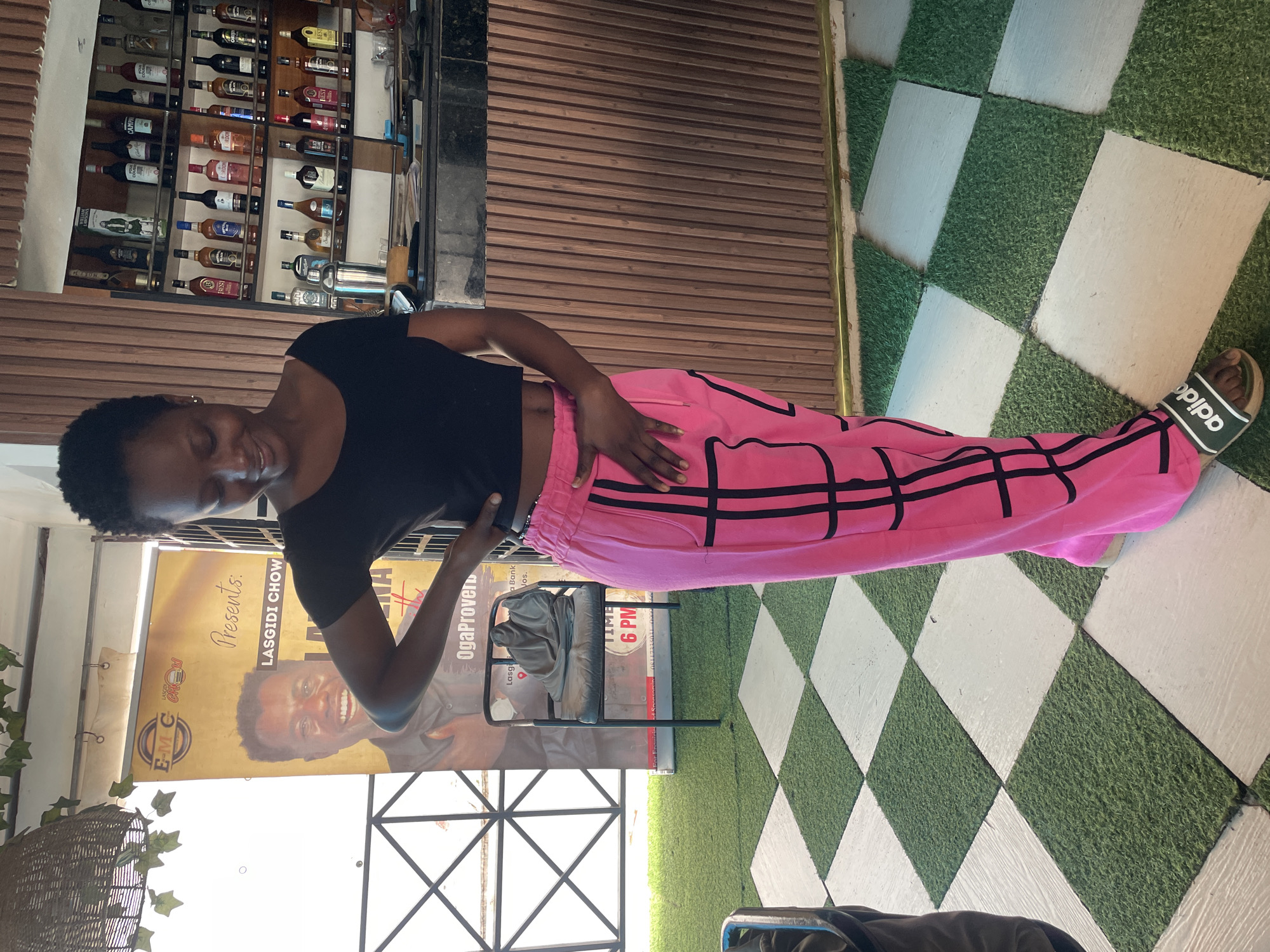 In Jos, Nigeria, a young Black woman with short, dark hair and a black crop top stands in front of a bar. She is wearing bright pink track pants with black stripes and Adidas slides. Her left hand is on her hip, and her right hand is resting on her pants. Behind her, a bar shelf is stocked with various bottles of liquor. The floor is a checkered pattern of green turf squares and white squares. A poster is visible to her left, featuring a man smiling and some text in English. A chair with a draped cloth sits in front of the poster.