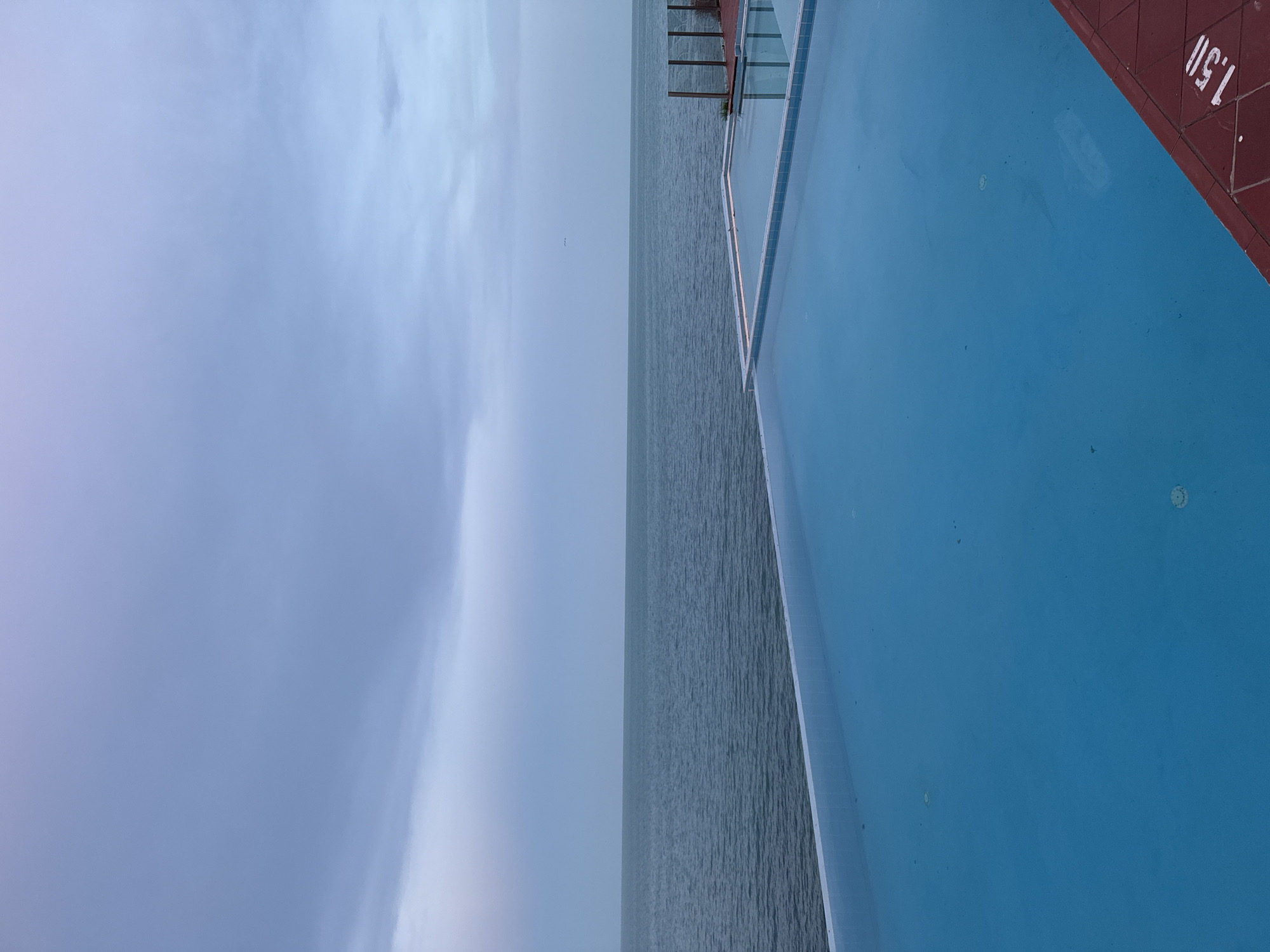 The image depicts an infinity pool overlooking a body of water, likely the Black Sea, given the Varna, Bulgaria context. The sky is overcast with a gradient of soft purples and blues, suggesting either dawn or dusk. The pool's surface is calm and reflects the muted tones of the sky. The edge of the pool is lined with white tiles, and a red tiled walkway is visible in the foreground with a depth marker "1.50". In the distance, a faint horizon separates the water from the sky. There are no people or animals visible in the frame. The scene is tranquil and captures a serene coastal atmosphere.