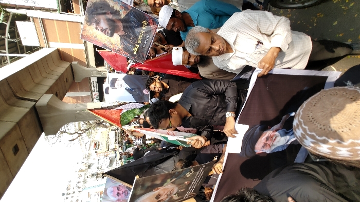 A dense crowd of individuals, primarily men of varying ages, is gathered on an urban street in Dhaka, Bangladesh, during daylight hours. The location is identifiable by a visible sign reading "বাংলাদেশ সচিবালয়" (Bangladesh Secretariat) in the background. An elevated concrete structure, characteristic of a metro line or flyover, spans overhead. Participants are engaged in what appears to be a public demonstration or procession, holding numerous posters and flags. Prominent posters feature portraits of Qassem Soleimani, Ayatollah Ali Khamenei, Yahya Sinwar, and another revered religious figure. A poster on the right identifies a bearded man with Bengali text as "শহীদ উসমান গনি" (Shaheed Usman Ghani). Flags include multiple black banners, a Palestinian flag, and various green, white, and red flags. Many individuals are wearing traditional Islamic headwear (topis). The scene is brightly lit by sunlight, revealing an urban environment with multi-story buildings and a paved road. A motorcycle is partially visible on the right periphery.