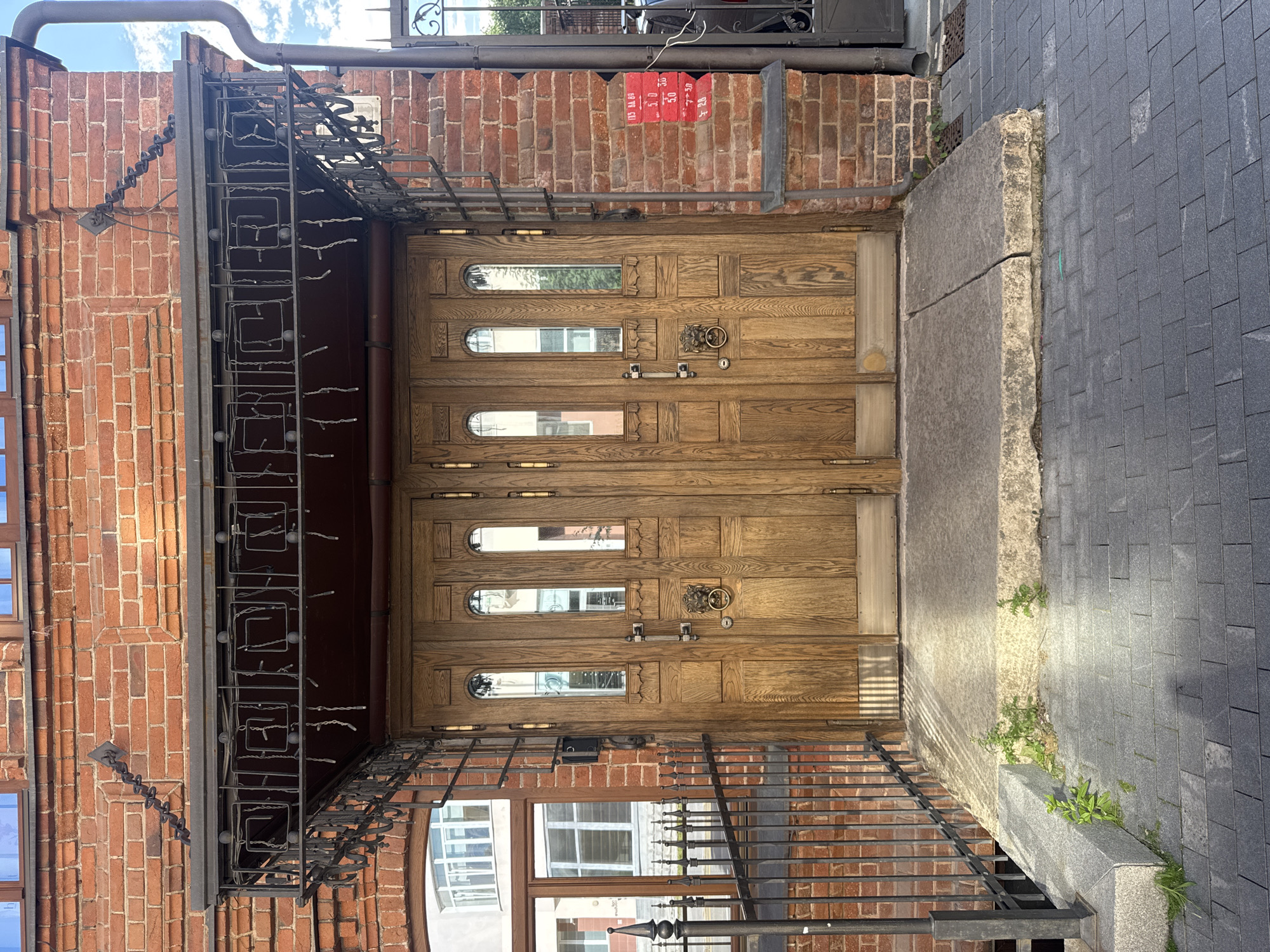 A vertical shot shows a double wooden door, likely the entrance to a building in Yekaterinburg, Russia. The doors are made of oak and feature several small, arched windows, some of which offer glimpses of the interior. A decorative lion's head door knocker is visible on the right door, along with two metal handles and a lock. Above the doors, a metal awning with intricate wrought-iron detailing is supported by vertical beams. To the left of the entrance, a brick wall is partially visible, with a light fixture attached to it. To the right of the doors, a walkway paved with dark gray rectangular tiles leads away from the entrance. A small patch of greenery grows between the tiles and a concrete planter. In the background, above the awning, a red sign with white text, possibly denoting prices or regulations, is affixed to the brick wall. The overall scene suggests a daytime setting under a bright sky, indicated by the natural light illuminating the pavement and casting shadows.