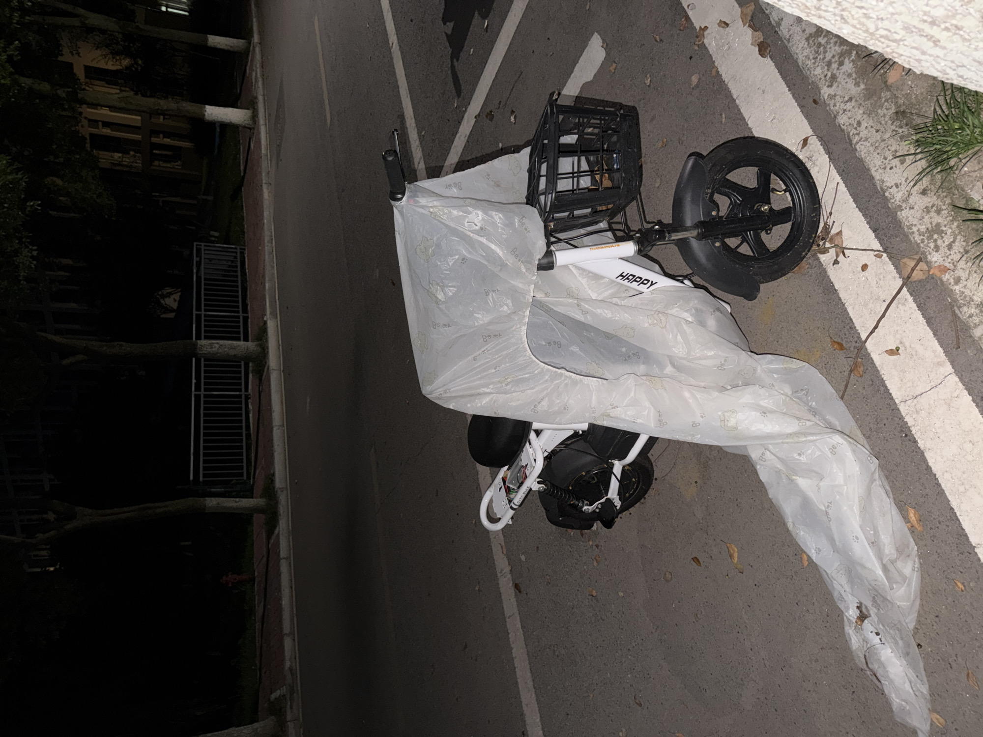 An electric bicycle, white with a black seat and basket, is parked on the side of a paved road at night. The bicycle is covered by a translucent white plastic sheet that extends onto the road. The plastic sheet appears to have a repeating pattern of small drawings. A black basket is attached to the front handlebars. The word "HAPPY" is printed in black on the bicycle's frame. The scene is set in a city environment, with trees lining the sidewalk and a metal fence visible in the background. The ground is asphalt, with fallen leaves scattered on the road and sidewalk. A white line marks the edge of the road. The lighting suggests artificial illumination, common in urban settings at night. The location is identified as Jiangjin, China.
