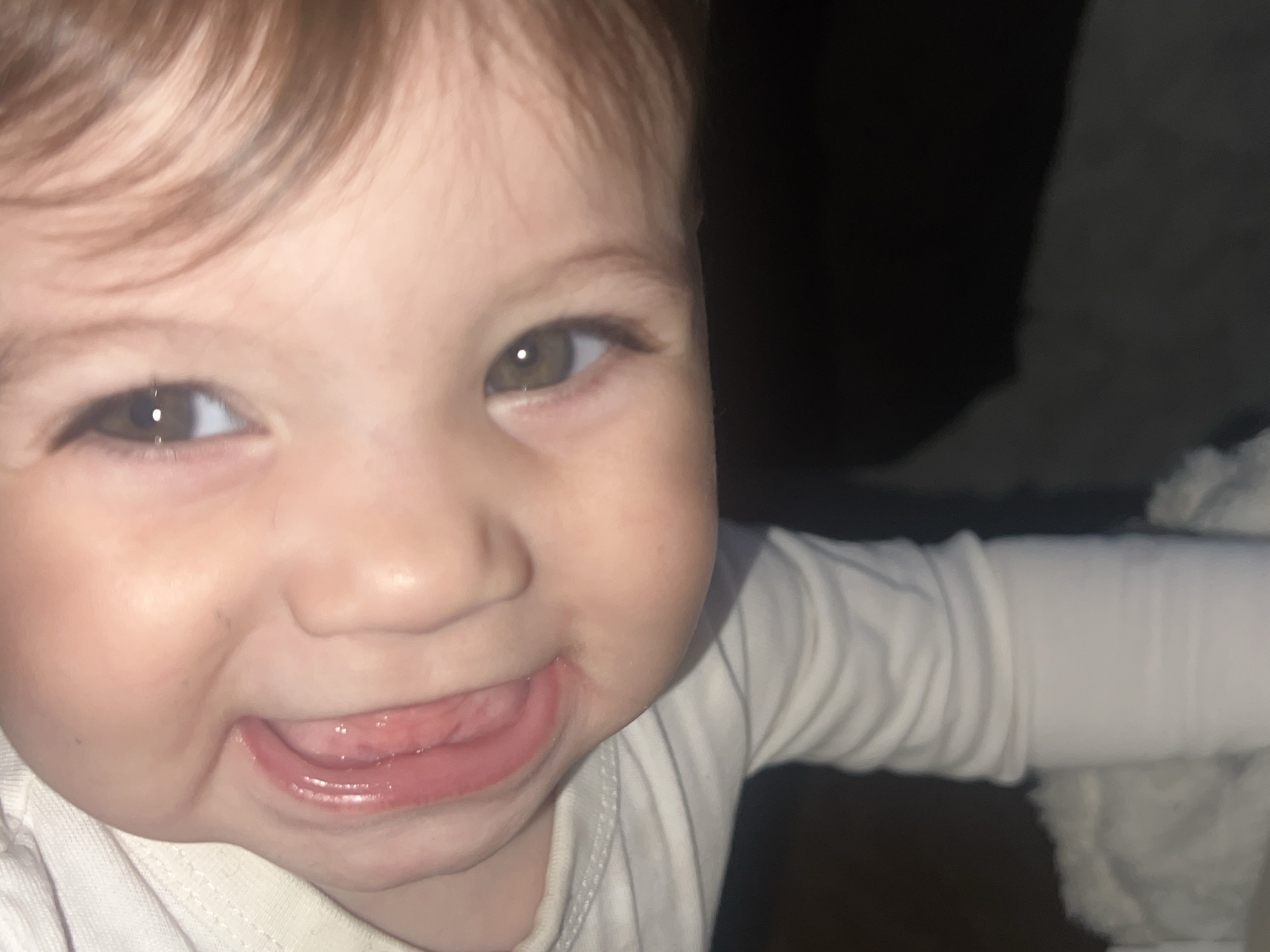 A close-up shot depicts a young child with light brown hair and brown eyes, smiling with their tongue slightly protruding. The child is wearing a light-colored, long-sleeved shirt. The background is dark and indistinct, with a hint of what appears to be a light-colored soft object on the right side of the frame. There is no indication of Elena, Bulgaria or any location or time.