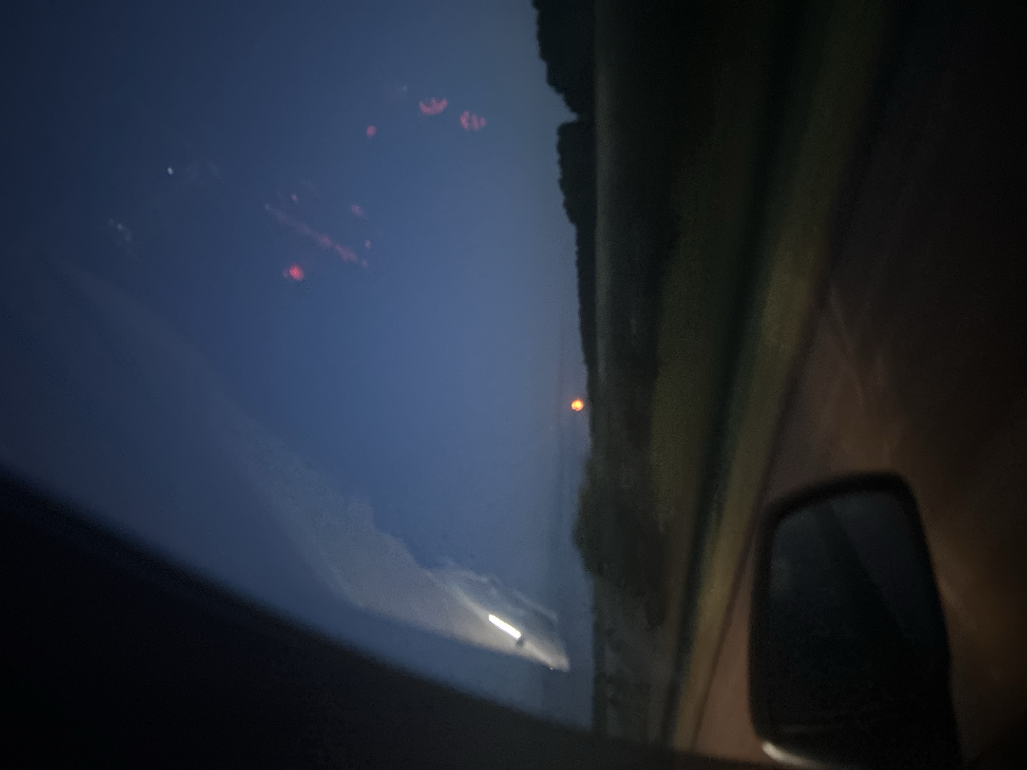 This media file captures a nighttime scene, likely from inside a vehicle, as suggested by the reflection on the windshield and the presence of a side mirror. The sky is a deep, dark blue, indicating it is either dusk or pre-dawn. There are several blurred, red lights in the sky, possibly distant aerial lights or reflections.
On the right side of the frame, a dark, silhouetted landscape with a defined edge runs vertically, suggesting trees or structures lining a road. A small, bright orange orb is visible in the distance against the dark landscape. The windshield reflects a bright streak of light, likely from oncoming headlights. The overall impression is one of travel through a rural area at night. The location context of Tyukalinsk, Russia, suggests a likely setting in a flat, open landscape typical of the region. No people are visible, and no text is present.
