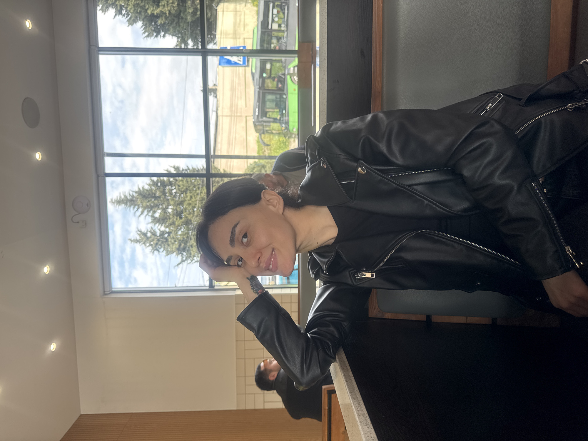 The image, taken in what appears to be a cafe or public space in Tbilisi, Georgia, features a young woman as its primary subject. She is shown from the chest up, leaning casually with her right arm bent, resting her head on her hand, and smiling towards the camera. She has dark hair, styled down, and prominent eyebrows. She is wearing a black leather jacket with visible zippers and a black top underneath. A tattoo can be seen on her right forearm, just above the wrist.
She is seated at a dark-colored table or counter, which dominates the lower right foreground. Behind her is a large, multi-paned window, offering a view of the outdoors. Through the window, green conifer trees are visible under a partly cloudy sky. A green public bus can be seen on a street or road outside, along with a blue rectangular road sign, though the specific symbol or text on it is not clearly decipherable.
In the background, further inside the establishment, another person (a man) is partially visible, seated at a distance. The interior ceiling is white, with several recessed spotlights and a circular speaker visible. The overall setting suggests a relaxed daytime atmosphere.