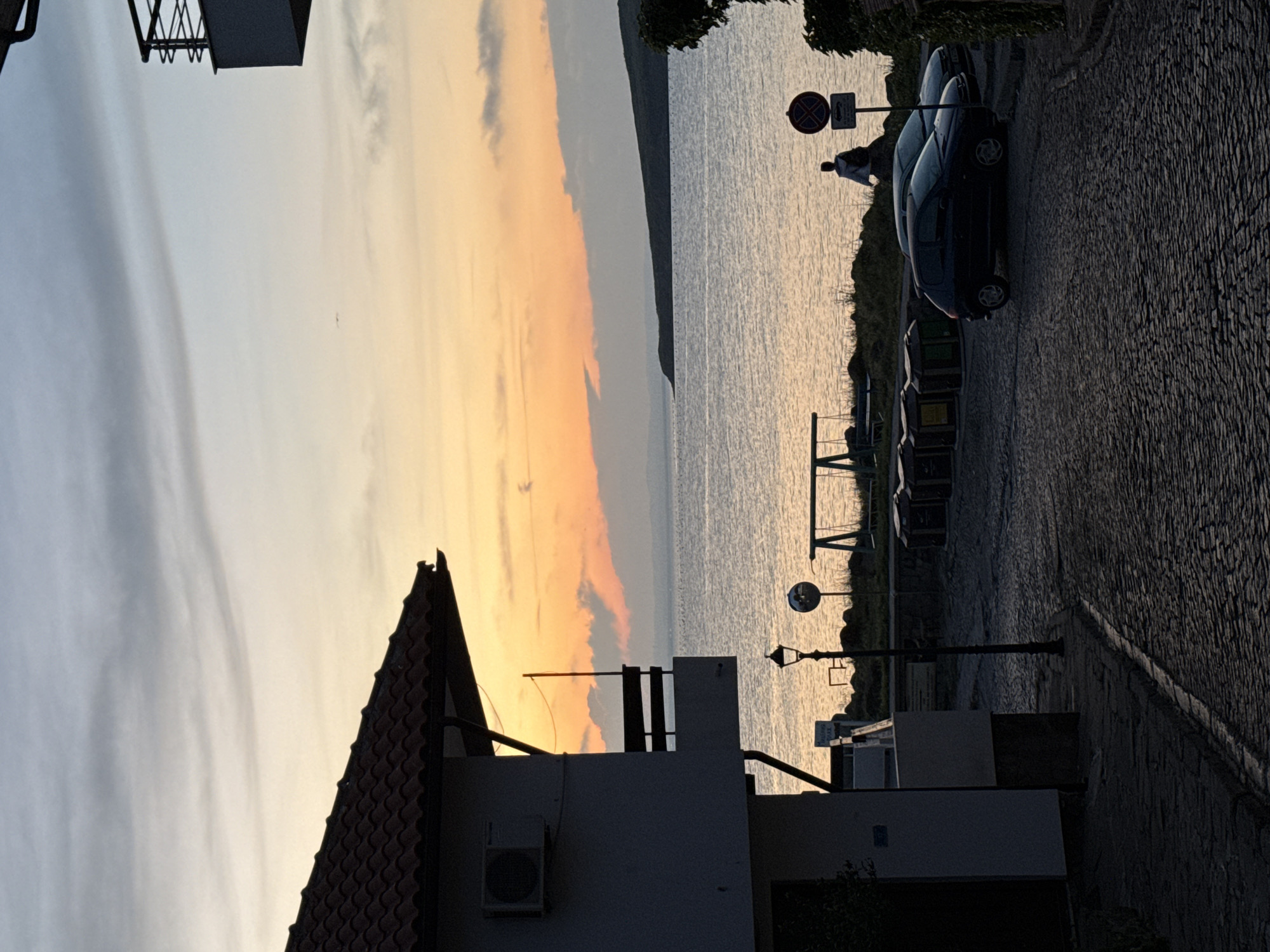 The image depicts a tranquil evening scene in Sozopol, Bulgaria, likely along the waterfront. The sky is painted with soft hues of orange and yellow from the setting sun, transitioning into pale blue and gray clouds. A calm body of water, possibly the Black Sea, stretches towards a distant, low-lying landmass.
In the foreground, a cobblestone street curves gently, leading towards the water. On the left, a portion of a building with a tiled roof and an air conditioning unit is visible. A street lamp stands to the right of the building. Further along the street, two dark-colored cars are parked. To the right of the street, a few trash bins are lined up. In the middle ground, a simple swing set is set up near the water's edge. A lone figure, silhouetted against the water, walks near a signpost that appears to indicate parking restrictions. The overall atmosphere is serene and peaceful, capturing the essence of a quiet evening in a coastal town.