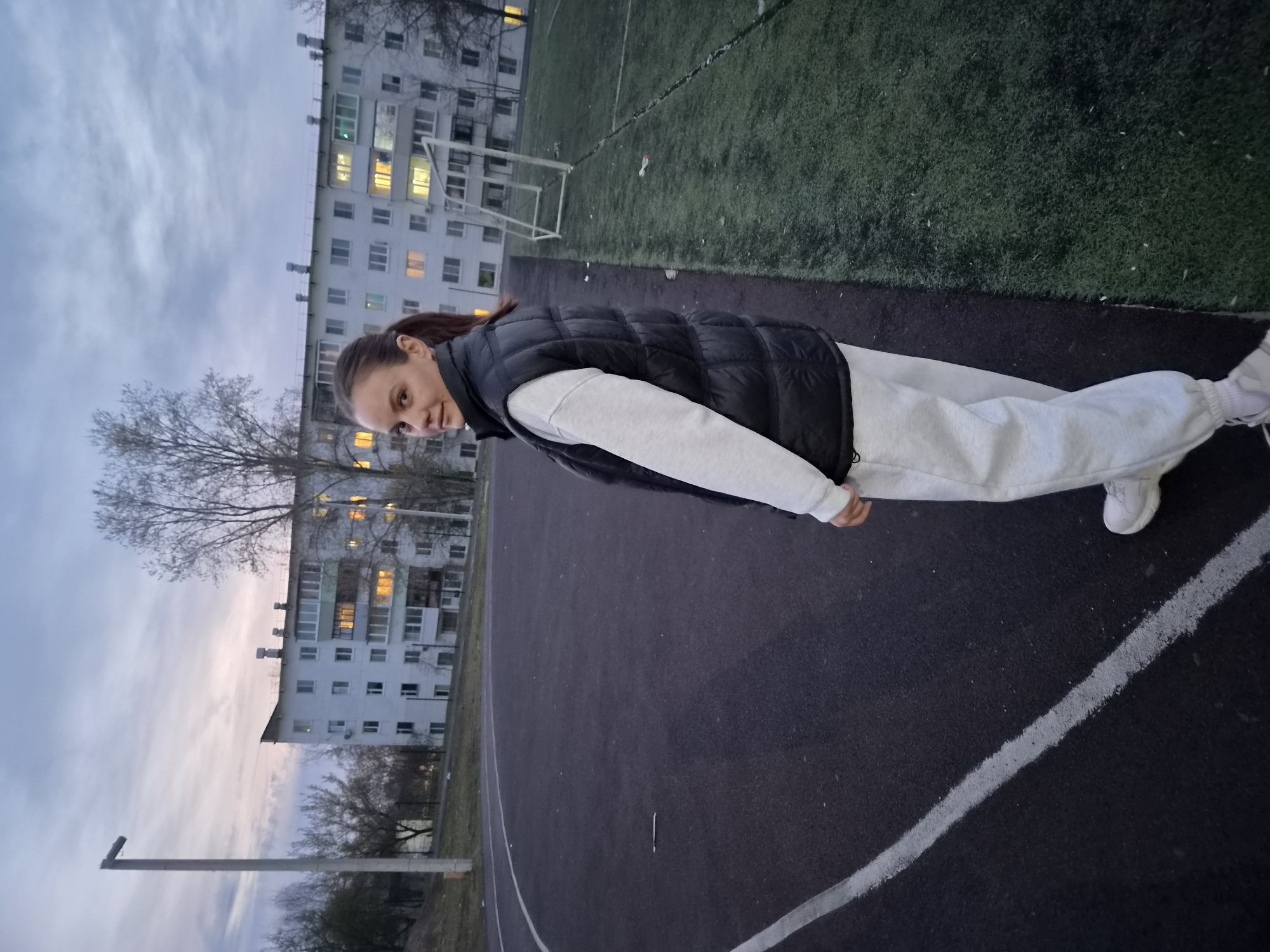 A young woman is captured in a plank position on an outdoor running track, likely performing an exercise. She is viewed from the side, looking towards the camera with a focused expression. Her dark hair is pulled back in a ponytail. She is dressed in a black sleeveless puffer vest over a light grey long-sleeved top, light grey sweatpants, and white sneakers.
The setting appears to be a sports ground or stadium located in Chaikovsky, Russia, possibly adjacent to a residential area. Behind her, to the left, is an artificial turf field, with part of a white soccer goal visible. Further in the background, multi-story apartment buildings with numerous windows are visible; some of these windows are lit up with warm, glowing light, suggesting it is either dusk or early evening. The sky above is overcast with soft, muted tones of blue and grey. Bare trees are scattered around the periphery of the buildings. The running track itself is dark asphalt with a white dividing line visible in the foreground. No discernible text is visible in the image.