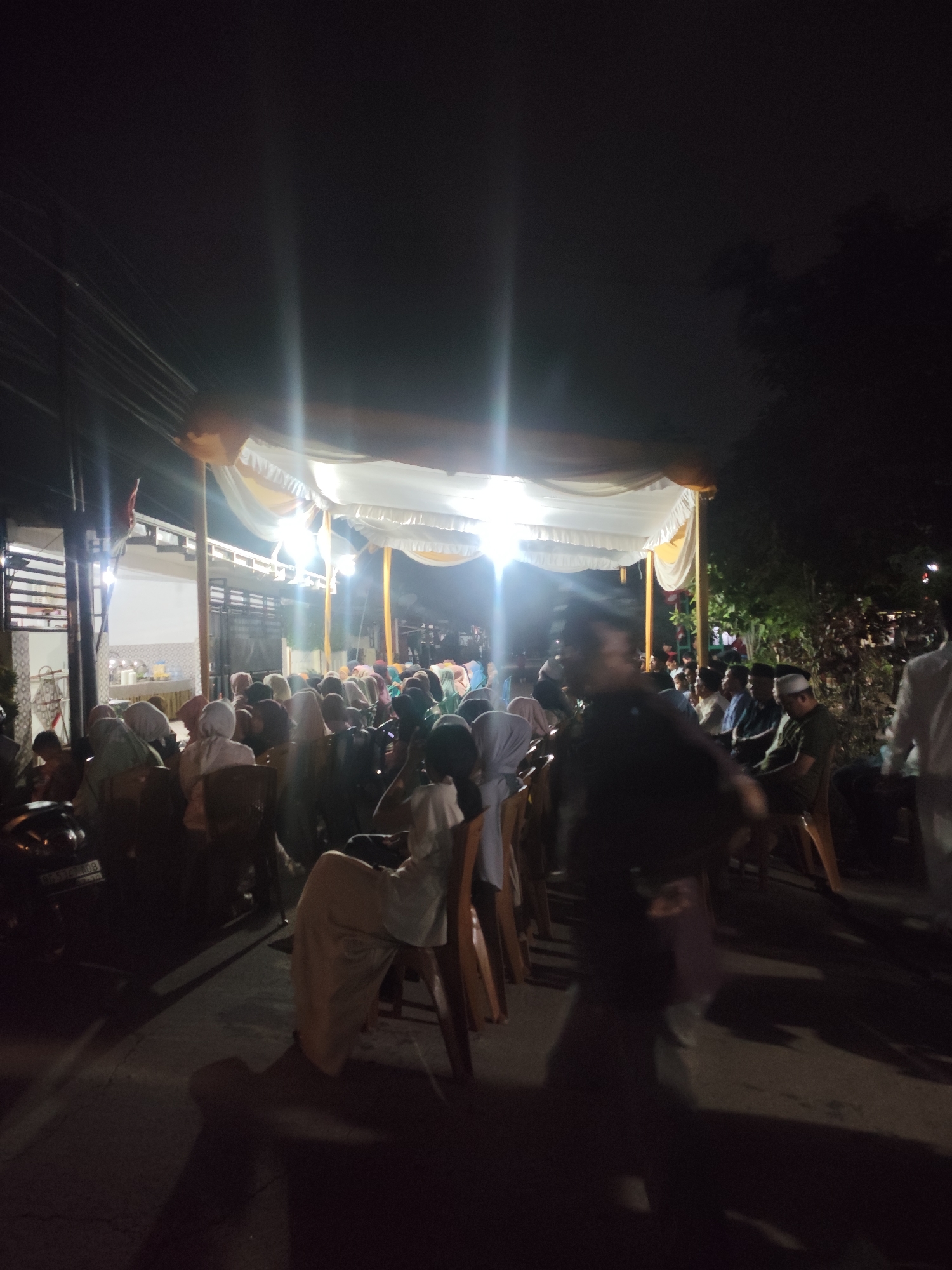 The image depicts an outdoor gathering at night, likely an event or ceremony, in Palembang, Indonesia. A white and yellow tent is erected, illuminated by bright lights that create lens flares. A crowd of people, many wearing headscarves, are seated on chairs facing the tent. Some individuals are standing or walking, and their faces are not clearly discernible due to the low light and distance. In the foreground, a person in a dark garment is walking, and their shadow is cast on the ground. A motorcycle is parked on the left side of the image. The environment appears to be a street or open area adjacent to buildings. The overall atmosphere is of a community event held after dark.
