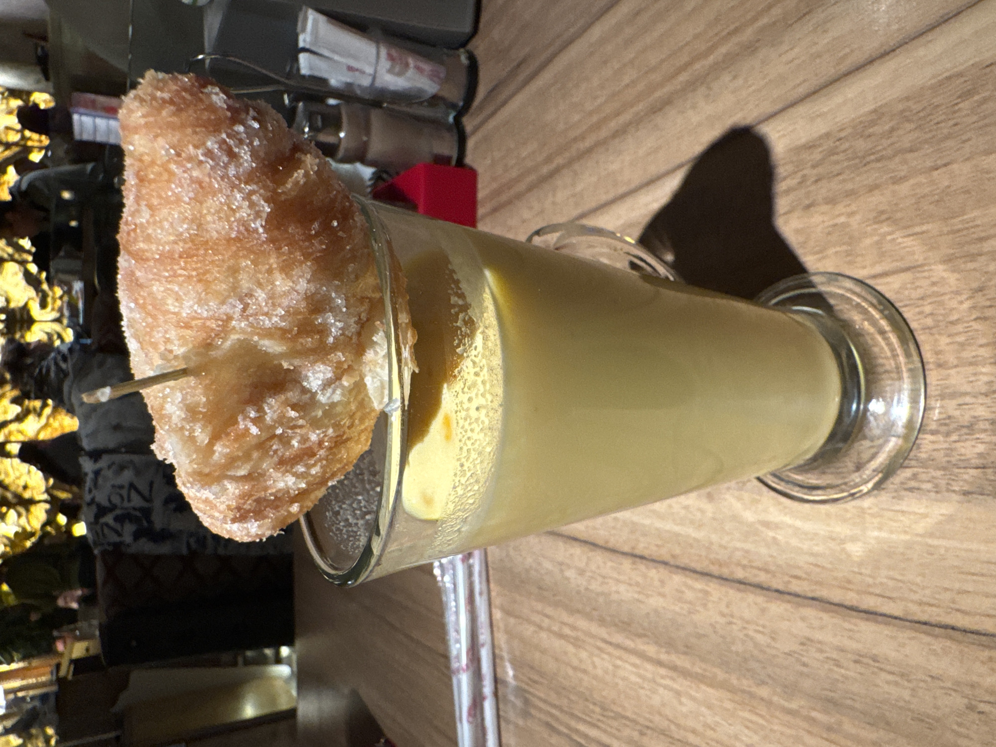 The image shows a tall glass of milky yellow drink, possibly horchata, topped with a pastry skewered with a stick. The pastry appears to be a croissant or similar, coated with granulated sugar. The glass rests on a wooden table. In the background, out of focus, are people sitting near a decorative golden branch or tree structure.
Given the presence of horchata and the background details, it's plausible that this photo was taken at a café or restaurant in Sofia, Bulgaria, possibly one with a modern or trendy interior design.