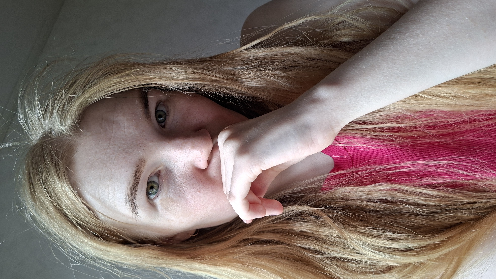 The image features a person, likely female, with fair skin, noticeable freckles across the nose and cheeks, and striking green eyes. Their long, wavy hair is a warm reddish-blonde color, spread around their head. The person is wearing a vibrant fuchsia or hot pink top or dress. One hand is partially covering the lower part of their face, specifically the mouth and chin area, with fingers slightly curled. The gaze is directed towards the camera, conveying a somewhat introspective or curious expression.
The scene appears to be an indoor setting, with the subject lying down on a light-colored surface, possibly a bed or floor. The background is indistinct and light-toned, suggesting natural, soft lighting. The overall activity is a relaxed pose for a photograph, possibly a selfie. There are no specific external location cues visible within the image itself. There is no visible text in the image. The provided location context is Большой Букор, Russia.