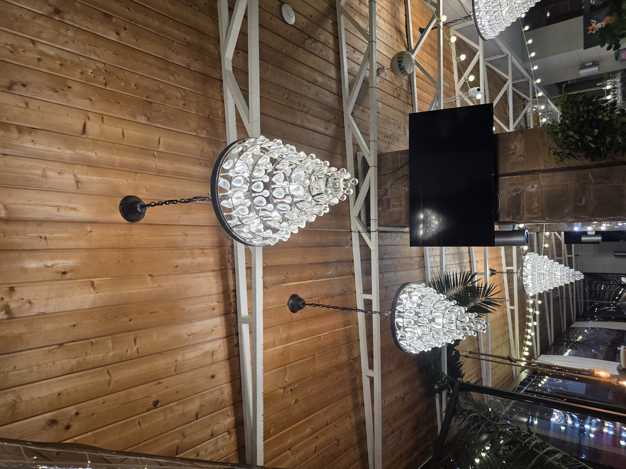 The image depicts an indoor scene with a wooden plank ceiling. Three crystal chandeliers are visible, two in the foreground and one further in the background. A large black flat-screen television is mounted on a stone-clad column. String lights with round bulbs are strung across the ceiling. Other elements include structural beams, a ceiling fan, and some foliage. The setting appears to be a restaurant or event space. The provided hints do not offer any additional information to determine the specific location in Sochi, Russia.