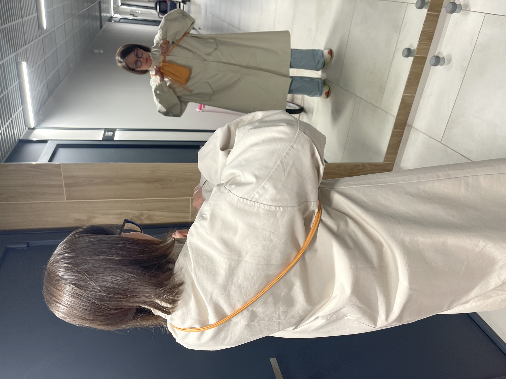 A person with brown hair and glasses is looking at their reflection in a mirror. The reflection shows a person wearing a long, beige trench coat and holding a small, orange purse. The person in the reflection is smiling and has their arms raised. The background shows a hallway with a tiled floor, a tiled ceiling with fluorescent lights, and a wooden paneling section on one wall. There is a door visible on the left and a row of cylindrical metal pegs on the right.