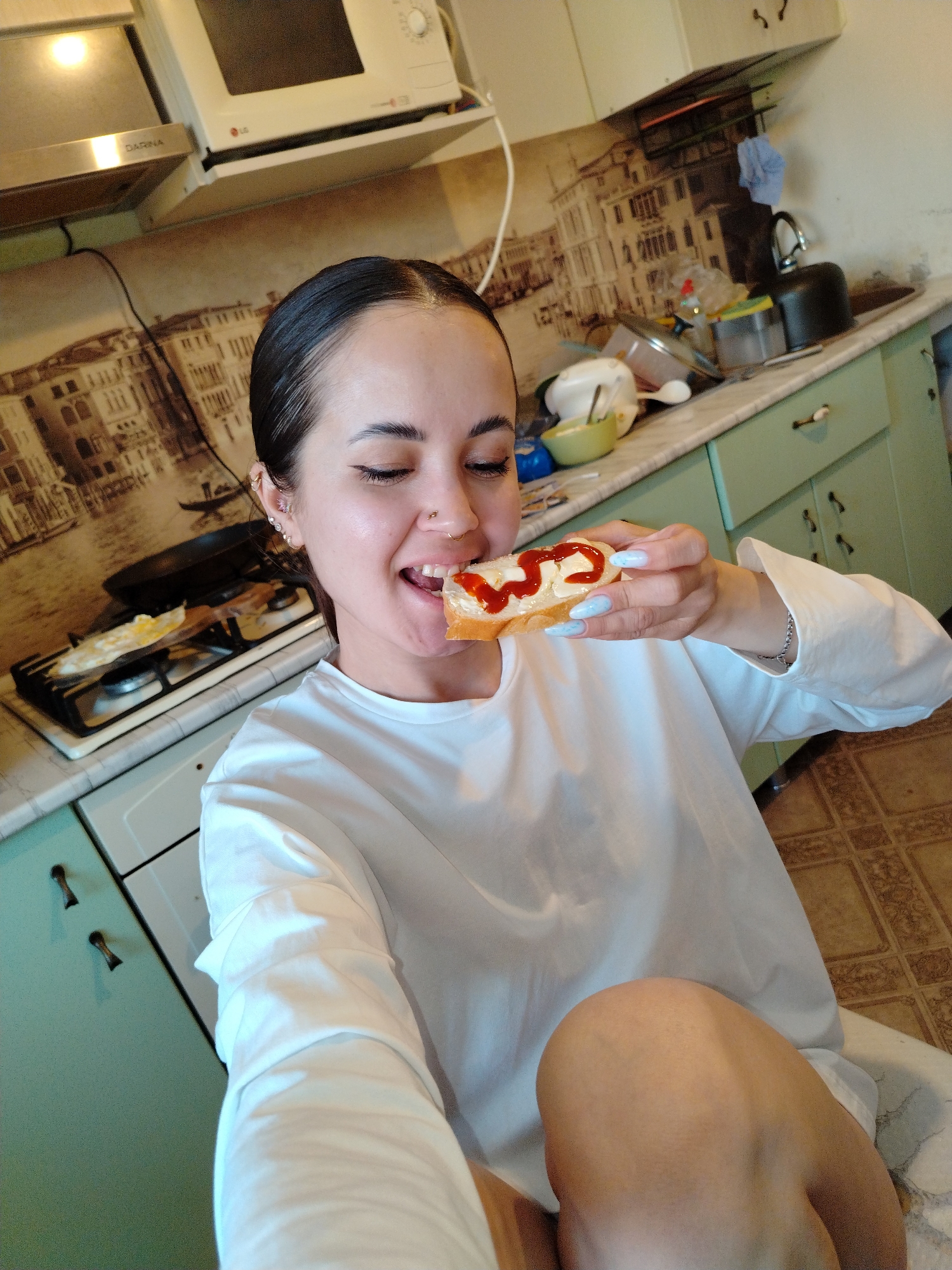 A woman is shown in a medium shot in what appears to be a kitchen. She is fair-skinned, with dark hair pulled back, and is wearing a white long-sleeved shirt. She has multiple piercings in her ear and a nose ring. She is smiling with her eyes closed and is holding a slice of bread with what looks like butter and a squiggle of ketchup on it, bringing it towards her mouth as if to take a bite. Her fingernails are painted a light blue color.
The kitchen has light green cabinets and a countertop that appears to be marble. In the background, there's a stove with a pan on it, a microwave mounted above the stove, and a backsplash with a mural of what looks like buildings. The area around the sink is cluttered with dishes and kitchen items. The floor has a tiled pattern. The city and location are unknown.