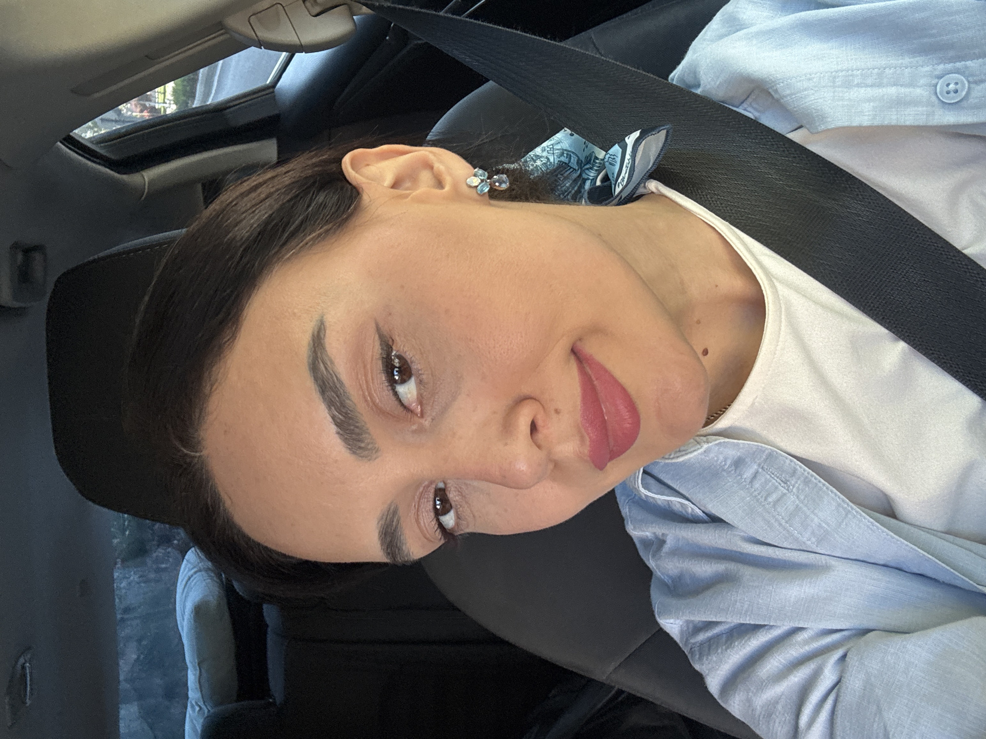 The image is a rotated selfie of a young woman taken inside a car. The woman is positioned with her head tilted to the left of the frame, looking slightly upward and to the left. She has dark hair, defined eyebrows, visible eyeliner, and pinkish-red lipstick. Her expression is relaxed, with a slight smile.
She is wearing a white top underneath a light blue, collared button-up shirt, which is unbuttoned and worn open. A dark seatbelt stretches diagonally across her chest and shoulder. In her right ear, she wears a small, light-colored floral-shaped stud earring. Behind her ear, a blue and white patterned fabric, possibly a scarf or hair tie, is visible. A thin gold chain is also visible around her neck.
The setting is the interior of a vehicle, indicated by the car seat's headrest behind her and the visible sunroof in the upper left background. Through the sunroof, natural light and blurred green foliage from outside are discernible, suggesting it is daytime. The car interior appears dark, primarily black, with a glimpse of a lighter seat in the far background on the left. No discernible text is visible on her clothing or in the immediate surroundings. The location context for the image is Tbilisi, Georgia.