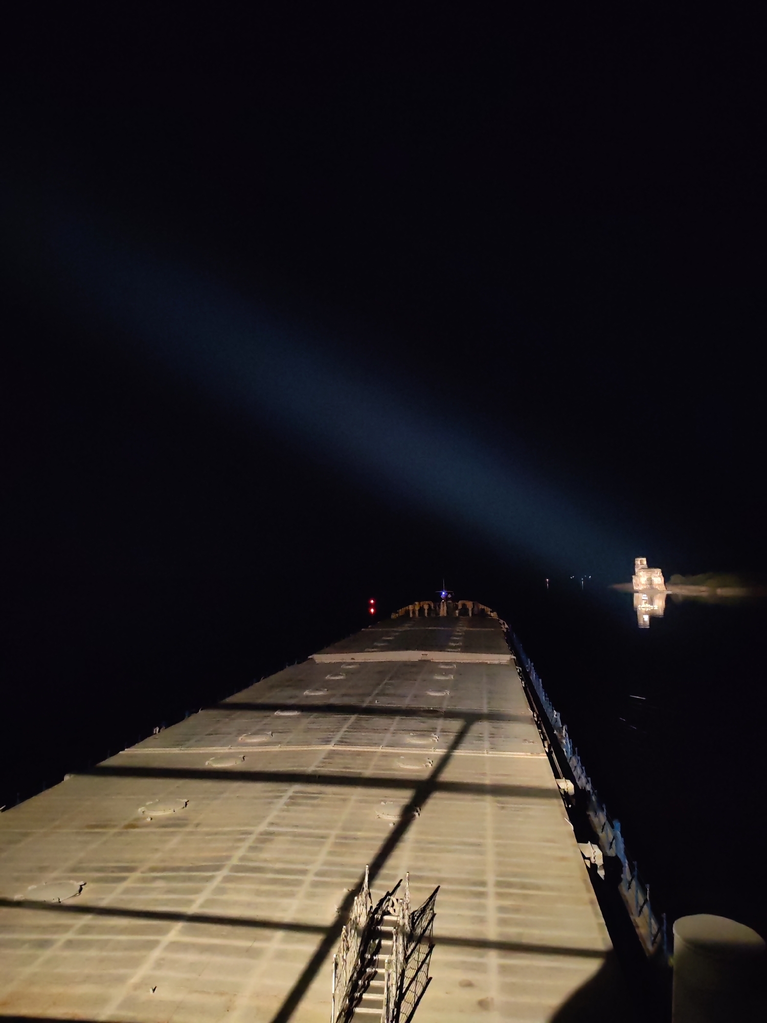 The image depicts the dark deck of a barge at night. The deck is illuminated by a spotlight. In the distance, there is a building or structure, possibly a ruined church, illuminated on the water and reflected on the surface. The location is likely the flooded area near the former village of Krokhino, Russia.