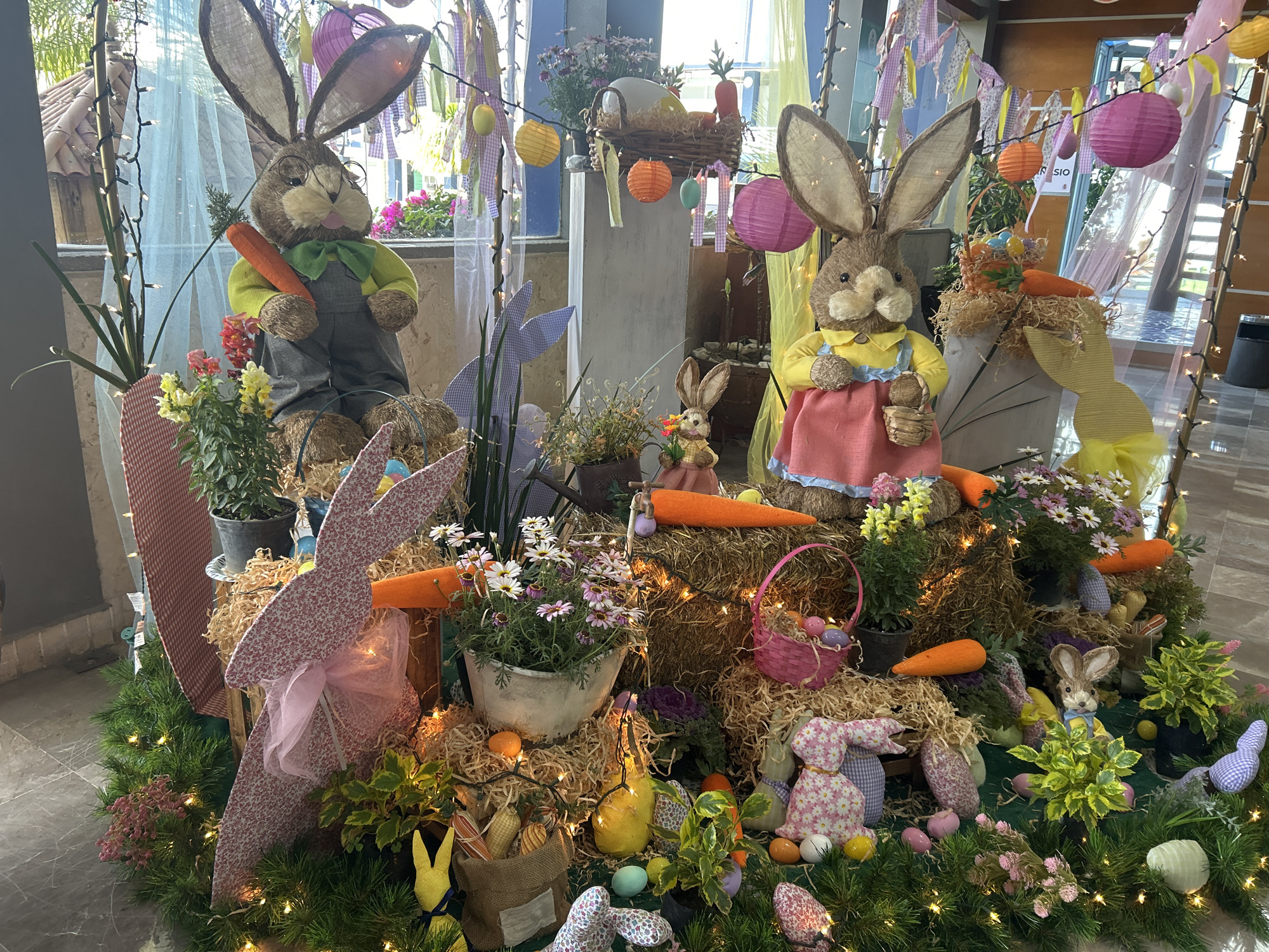 The image depicts an elaborate Easter display, likely located in a public space in Puebla, Mexico. The display features two large, stuffed bunny figures dressed in human clothing, positioned amidst a variety of Easter-themed decorations. These include artificial grass, hay bales, baskets filled with colorful eggs, carrots, assorted flowers, and smaller bunny figurines. Decorative lanterns and strings of lights are also present, enhancing the festive atmosphere. The background suggests the display is set indoors, possibly in a shopping center or hotel lobby. Architectural details visible through the window hint at the colonial style prevalent in Puebla. No people are visible in the shot.