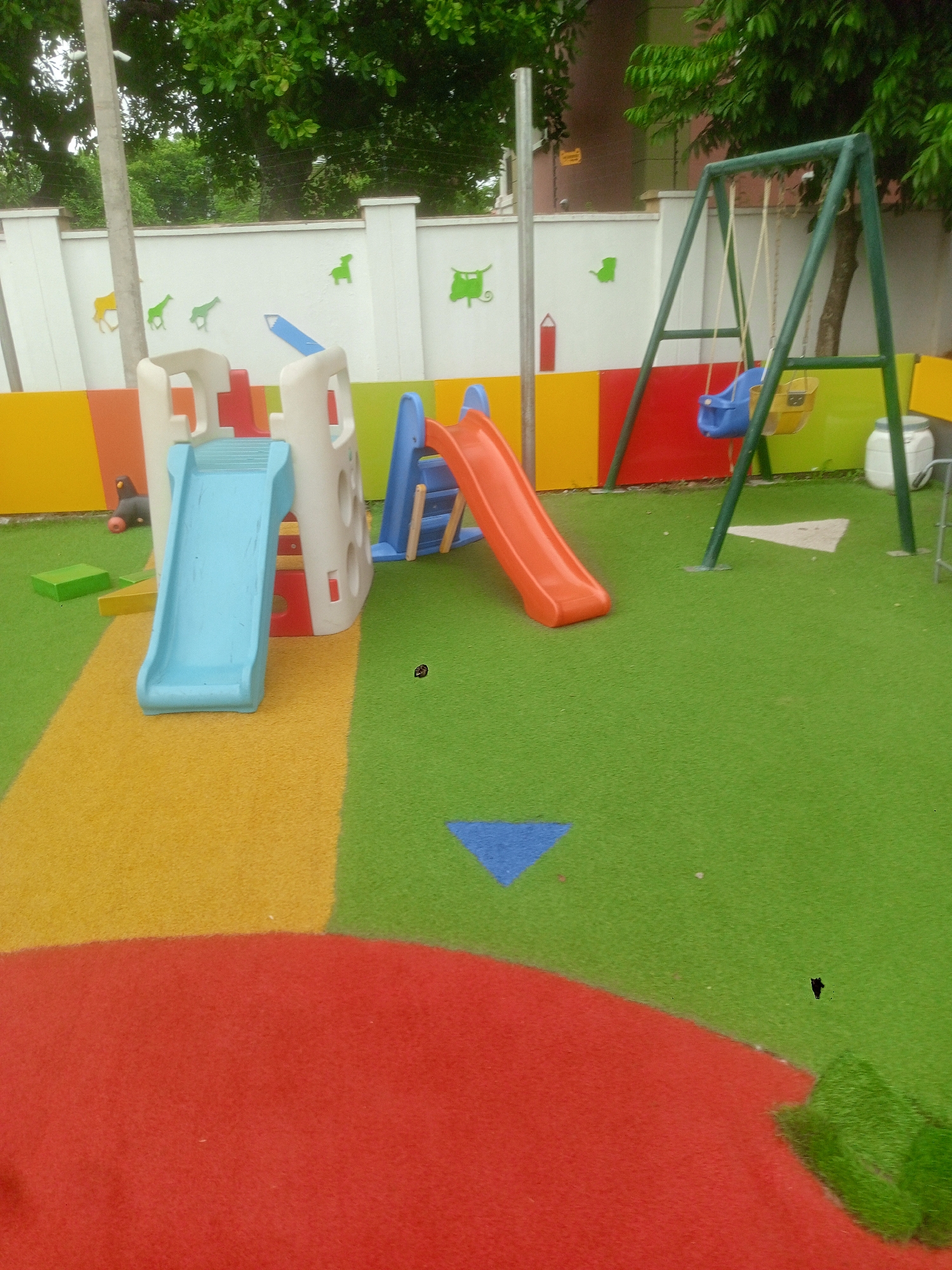 The image shows a brightly colored children's playground. There are slides, a swing set, and various shapes and colors on the ground. The playground is located outdoors, near a white wall with animal silhouettes. This scene appears to be located in Abuja, Nigeria.