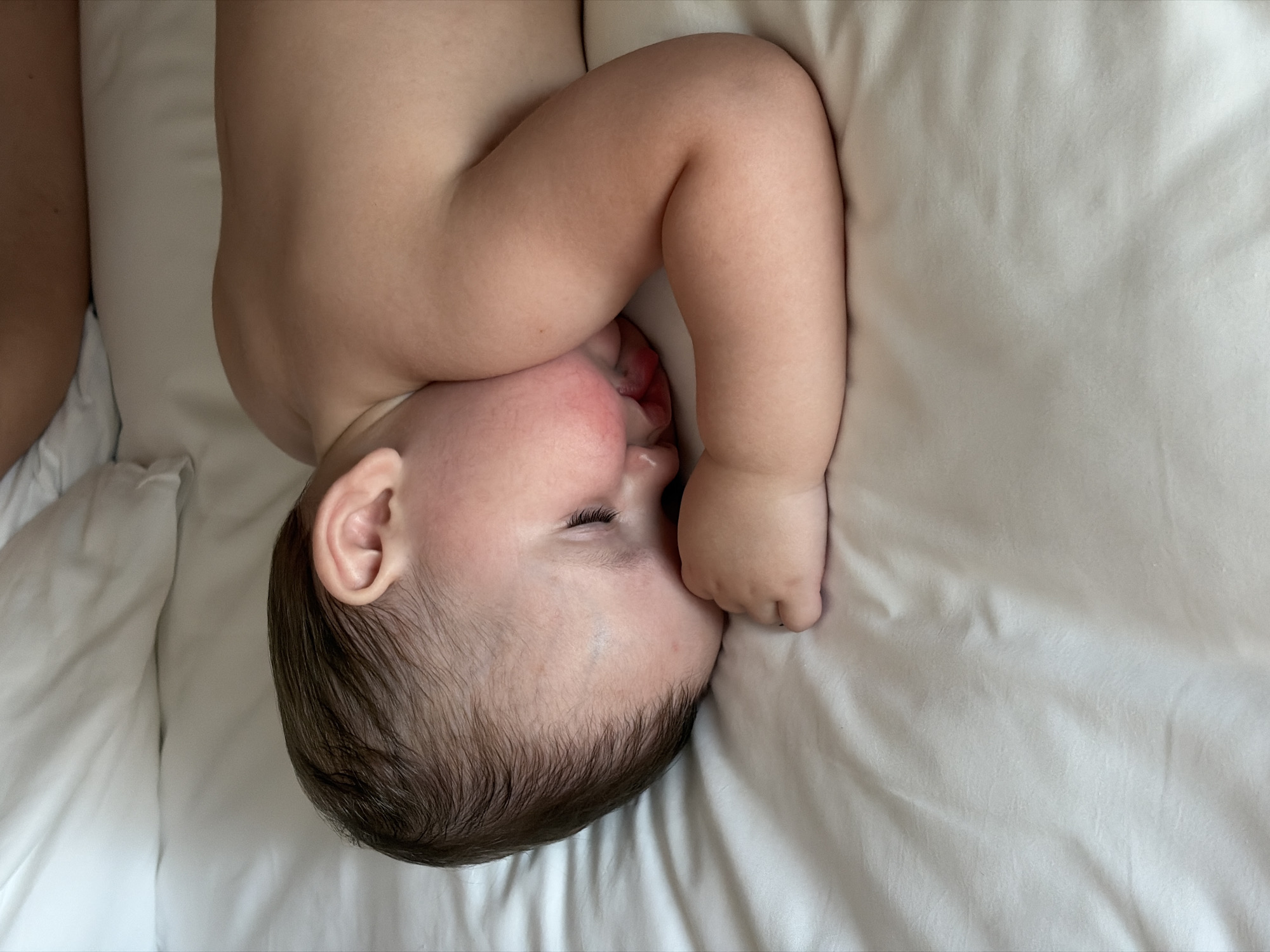A baby is lying on a white sheet, with their head turned to the side and their hand near their mouth. The location is not discernible from the image.