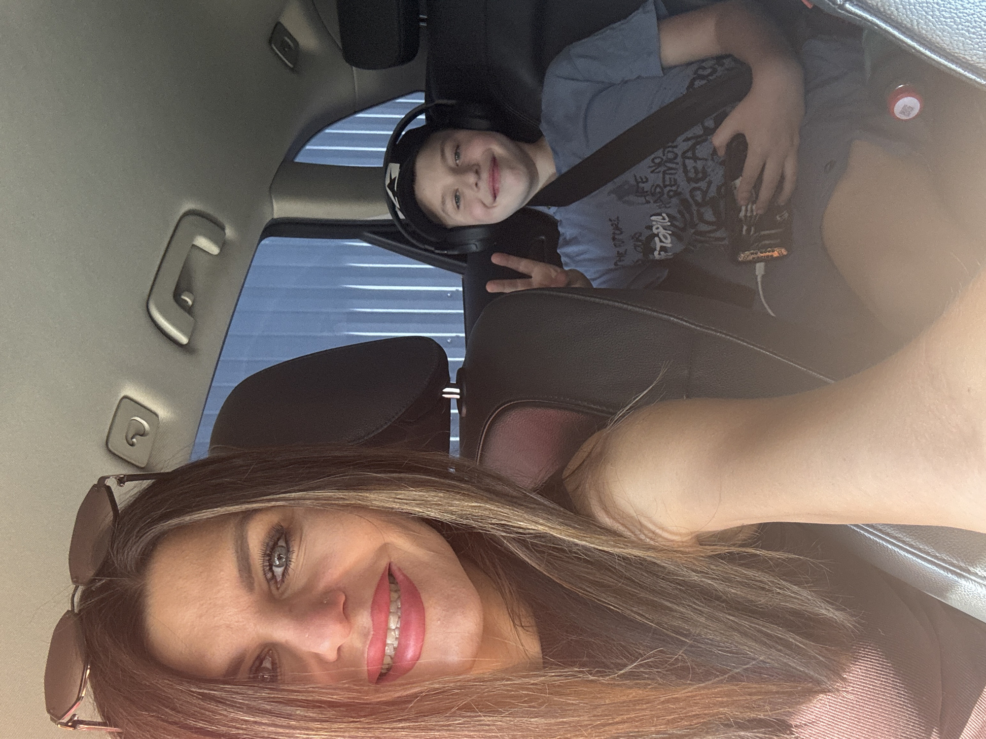 The photograph depicts two individuals in a car interior. The foreground features a woman with long, highlighted brown hair, wearing sunglasses perched on her head. She is smiling, revealing her teeth. In the background, a younger boy wearing headphones is seen in the back seat, giving a peace sign. He is wearing a seatbelt and holding what appears to be a handheld device. The environment appears to be the inside of a car, with the window showing what appears to be a structure with horizontal lines. The location in the city of Chaikovsky, Russia, is not evident from the image.