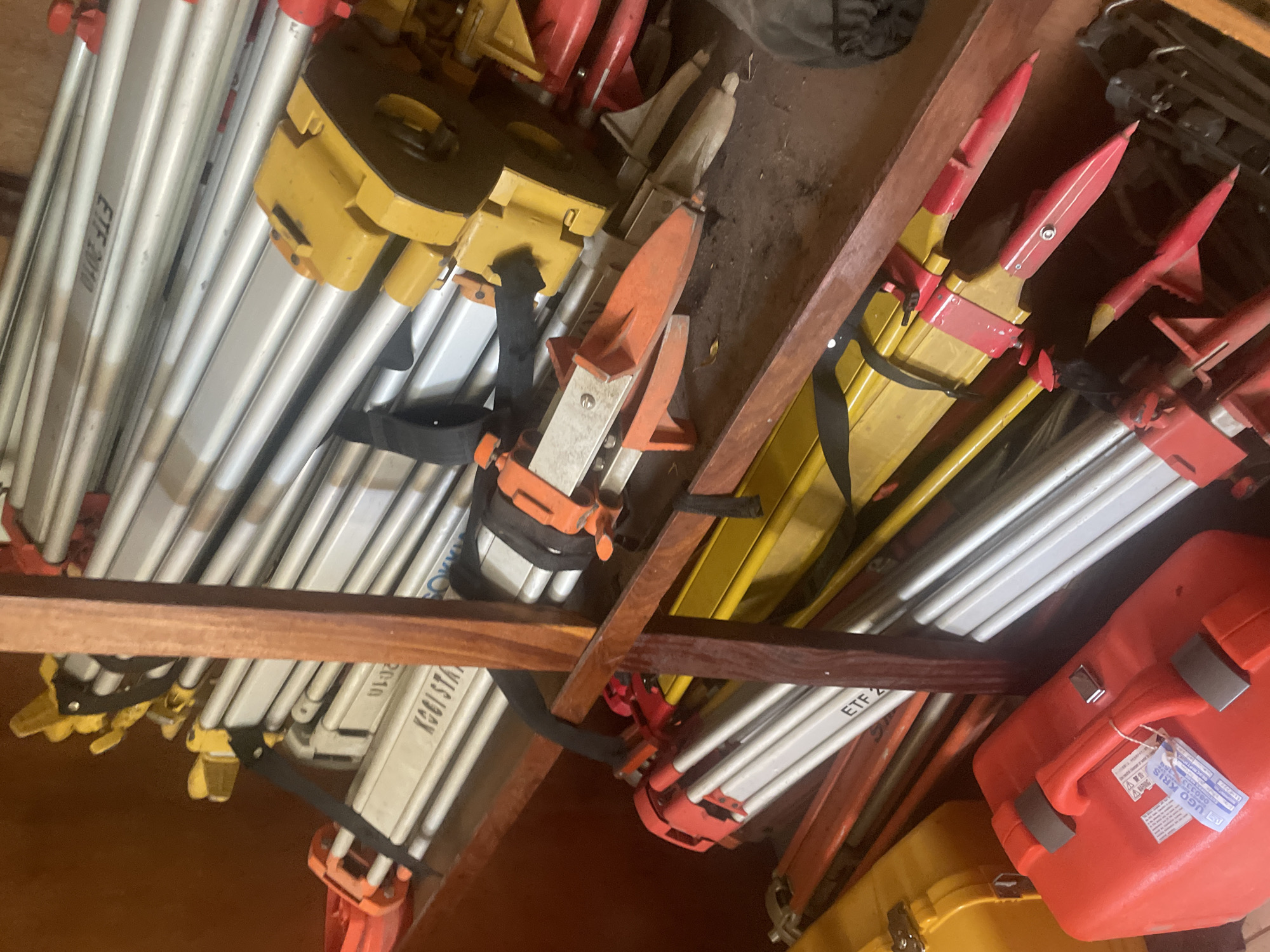 The image shows a close-up, top-down view of a collection of surveying tripods and their cases. The tripods are predominantly silver with yellow and red accents. Several are bundled together with black straps. Some of the tripod heads are yellow, while others are red. Visible text on the tripods includes "ETF 2000" and "KGSISTAH 20-10".
In the foreground and to the right, there are two bright orange cases, one of which is open, revealing a yellow interior. A label is attached to the handle of the orange case, but the text is largely illegible, though "UGD KRIS" and "030333" are partially visible. A yellow case is also visible beneath the orange one.
The scene appears to be an indoor storage area, possibly a workshop or a storage room, characterized by rough, dark surfaces and wooden shelving. The lighting is dim, suggesting an indoor environment possibly lit by artificial light. There are no people or animals visible in the image. The overall impression is one of organized storage of professional equipment. The location context provided is Lokoja, Nigeria.
