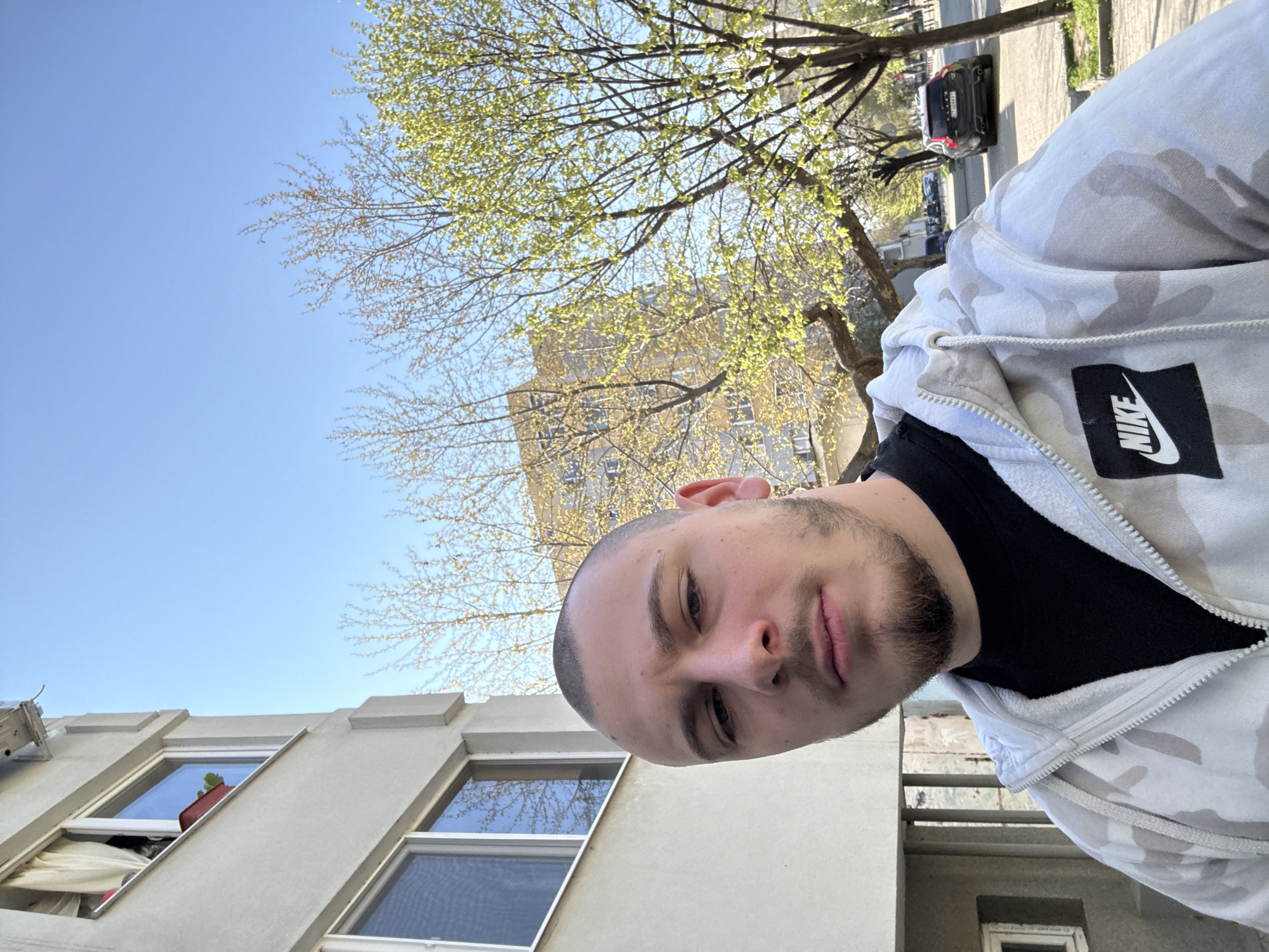 A medium-shot, slightly angled selfie depicts a young man wearing a light grey camouflage Nike hoodie over a black t-shirt. He has a shaved head and a beard. Behind him, a clear blue sky is visible, punctuated by a tree with budding green leaves. A beige building with windows is on the left of the frame. Further in the background, another building can be discerned through the tree branches. A street with parked cars can be seen in the far background. Based on the architectural style and the environment, it is likely in an urban setting such as the city of Stara Zagora, Bulgaria.