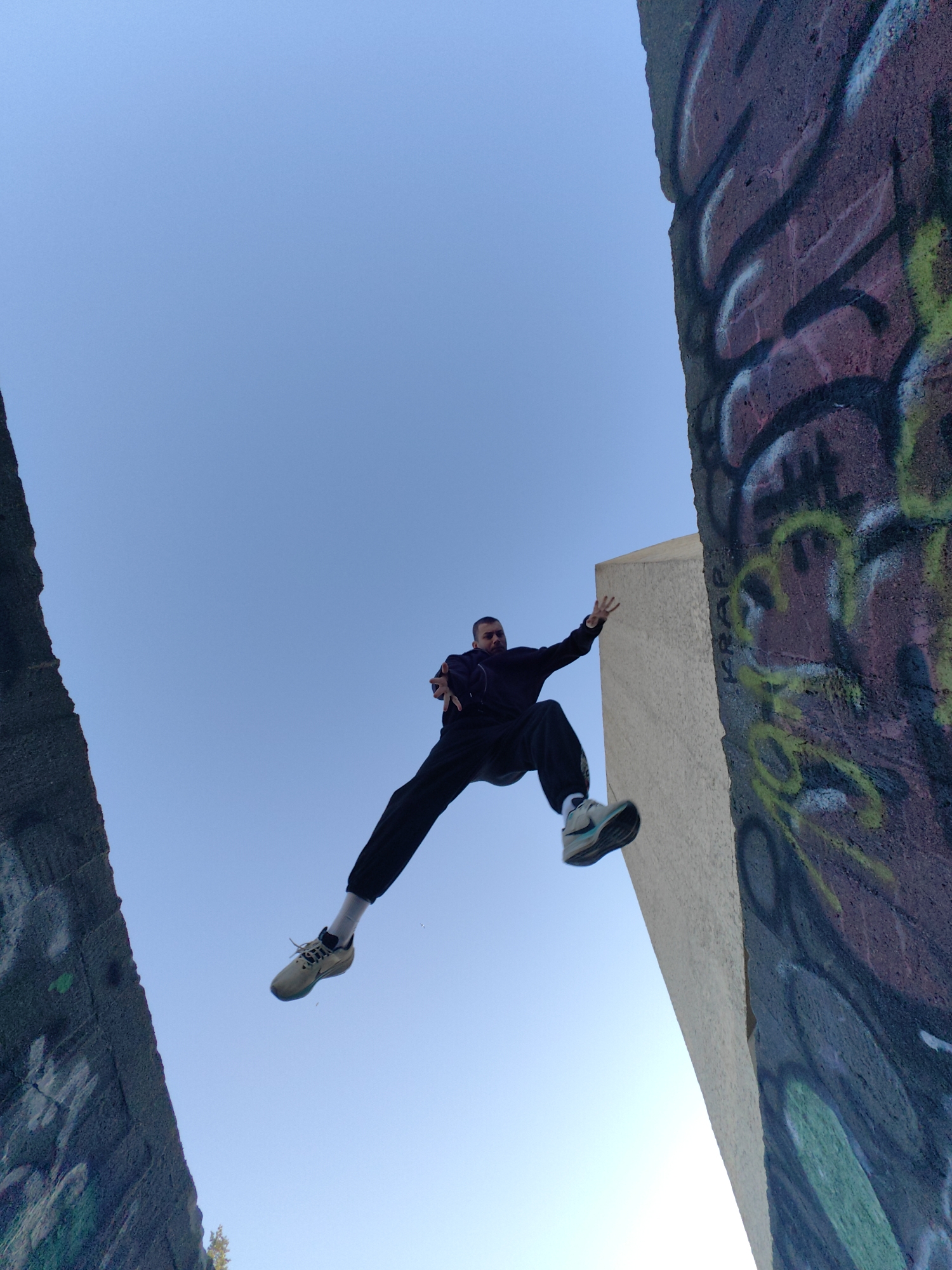 A dynamic, low-angle shot captures a male figure suspended in mid-air against a clear, bright blue sky. The man is positioned between two tall, rugged concrete structures, with his body stretched out in a jump or controlled fall. He wears a dark hoodie, dark pants, and light-colored athletic shoes featuring a distinct blue sole. His arms are outstretched, one hand slightly open, adding to the sense of movement and balance.
The concrete wall on the right is extensively covered in vibrant graffiti, showcasing bold patterns in purple, green, black, and white. The word "KRAP" is visibly written vertically in black on this wall. A lighter, unpainted concrete structure is visible behind the man on the right side. The concrete wall on the left is darker and appears to have some faint markings. The bright, uniform sky and clear visibility indicate daytime with good weather. The setting, located in Varna, Bulgaria, suggests an urban or industrial environment, possibly popular for activities like parkour or free running.