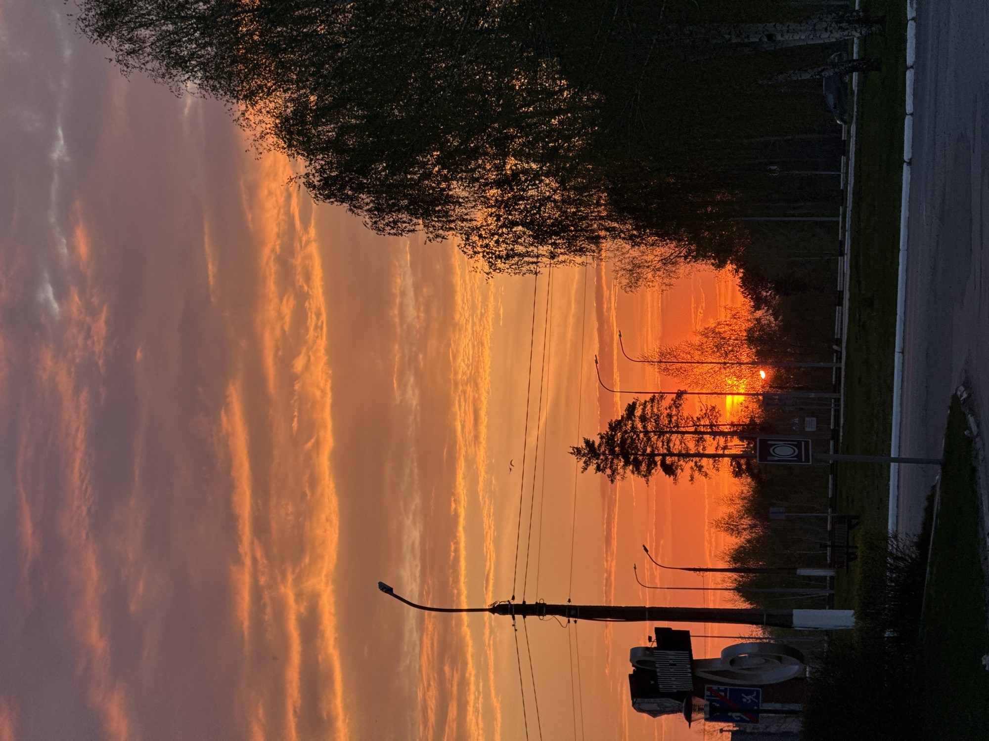The image shows a sunset over a street. The sky is filled with orange and pink clouds. Silhouetted trees are visible along the road. Streetlights line the street. There is a sign with symbols. Overhead power lines are also visible.
