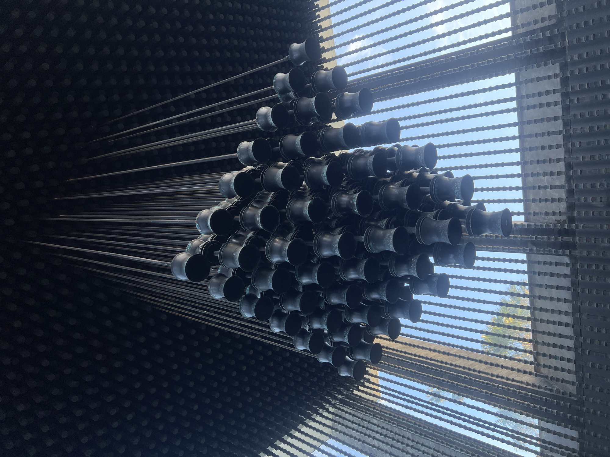 The image displays a large, complex art installation or architectural feature, likely indoors or in a covered space. The central element is a dense, dark cluster of numerous trumpet-shaped or bell-mouthed metal objects, stacked and interconnected, forming a roughly triangular or diamond-like mass.
From this central cluster, many long, slender metal rods or bars extend outwards. To the left, these rods lead towards a dark wall with a subtle, repeating pattern of small, lighter circular dimples. To the right, other rods extend towards a large rectangular opening or window. This opening is spanned horizontally by numerous textured bars or rods, which cast shadows and reveal segments of bright blue sky beyond, suggesting clear daylight. Some foliage, possibly palm trees, is faintly visible against the sky, consistent with the Kuta Selatan, Indonesia location.
The installation is predominantly dark gray and black, providing a strong contrast with the bright natural light filtering in from the right. The trumpet-shaped objects and the extending rods exhibit a metallic sheen, while the horizontal bars on the right show a distinct corrugated or segmented texture. No human activity or visible text is present; the image focuses solely on the static, intricate design of the structure. The overall impression is one of modern, industrial aesthetic.