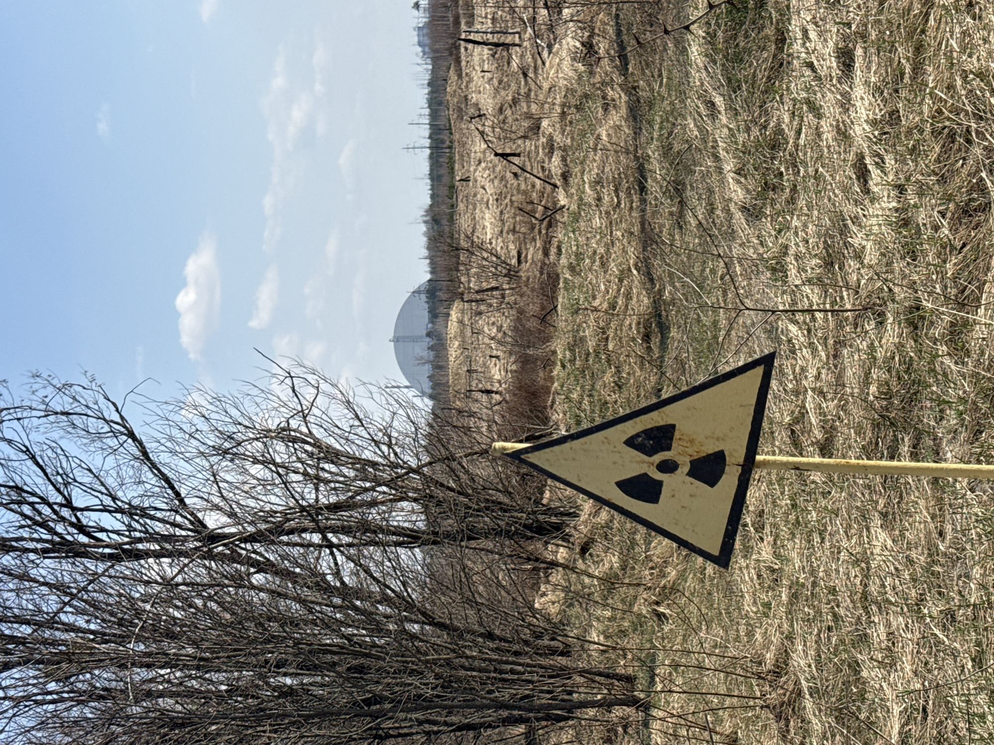 The image shows a landscape in what appears to be a rural area, likely near Хутор, Ukraine. A triangular radiation hazard sign is prominently displayed in the foreground. Behind the sign is a landscape of dry, grassy fields interspersed with bare trees and skeletal branches. In the mid-distance, a large, dome-shaped structure is visible, possibly associated with a power plant. The sky is light blue with some scattered clouds. The general impression is one of an abandoned or restricted zone.