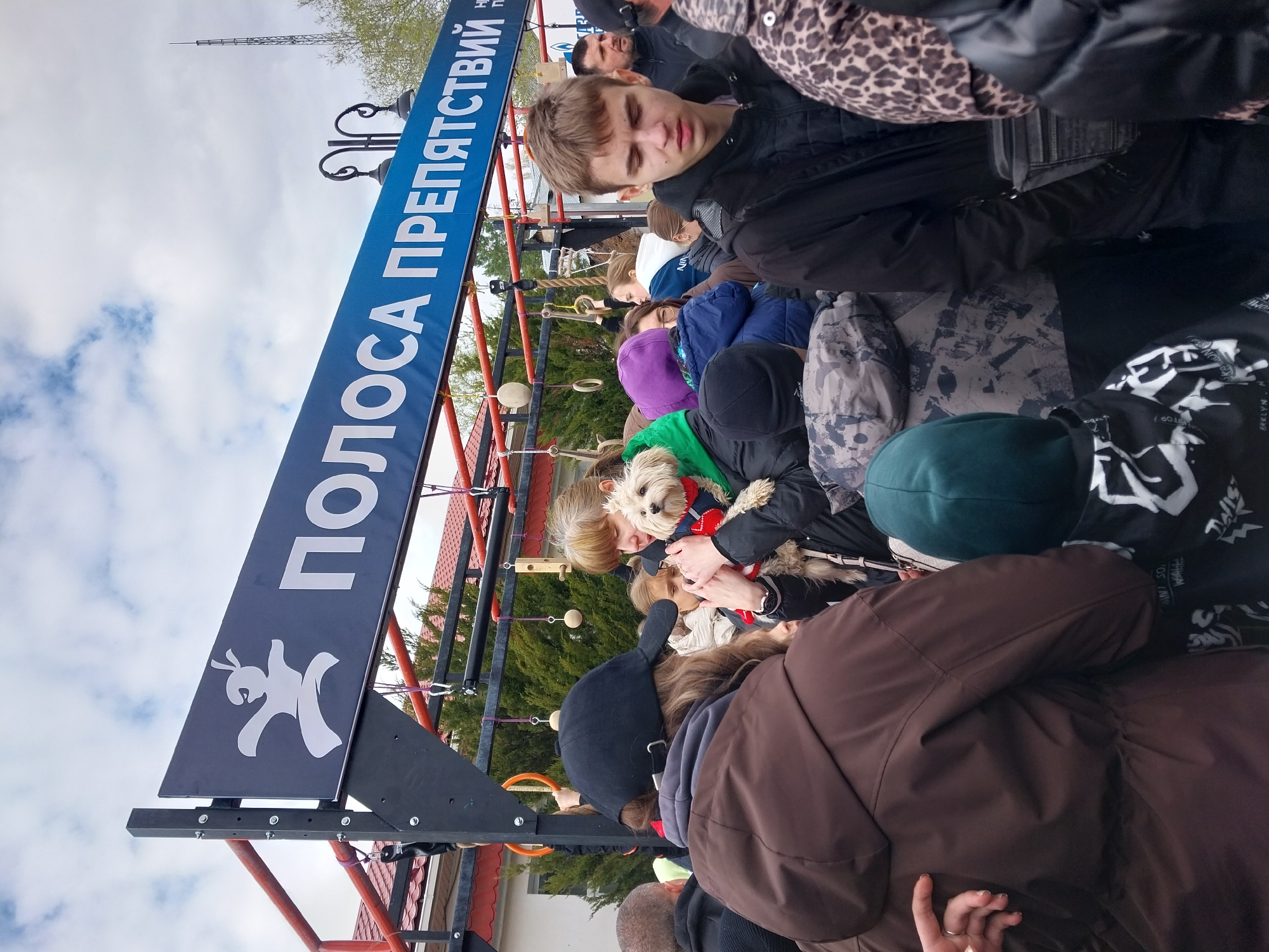 A crowd of people gathers around an outdoor obstacle course in Orenburg, Russia. The course, identified by a sign reading "ПОЛОСА ПРЕПЯТСТВИЙ" (Obstacle Course), features various climbing and agility challenges. Participants and onlookers wear jackets and hats, suggesting a cool climate. A woman in a green shirt holds a small white dog wearing a red and blue scarf.