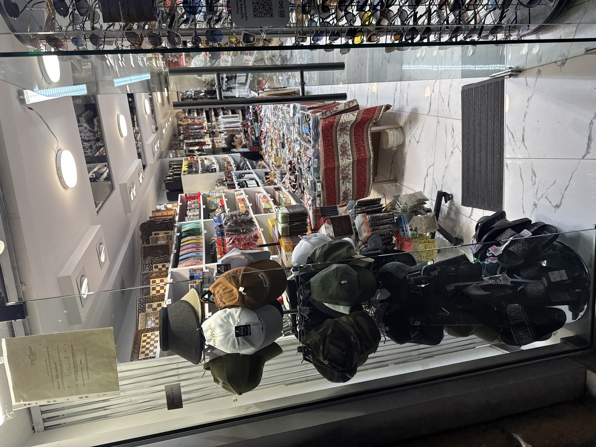 The image shows the interior of a small shop, likely a souvenir or accessory store, viewed through a glass door. The shop is located on the ground floor of a building in Yerevan, Armenia, as indicated by the Armenian text on a sign posted on the glass door.
Inside the shop, there are shelves filled with various items, including hats, sunglasses, scarves, and other accessories. The store appears to be well-stocked, with merchandise displayed on racks, shelves, and tables. In the background, a young man is seen attending to customers or organizing merchandise behind a counter. The flooring is made of white marble tiles. The lighting in the shop is provided by ceiling lights, and natural light enters through the glass door.
The foreground of the image features a display of hats on a tiered rack. These hats include baseball caps and visors in various colors and patterns. To the right, a rack filled with sunglasses is visible, showcasing a wide selection of styles. A dark gray doormat is placed on the marble floor in front of the glass door.