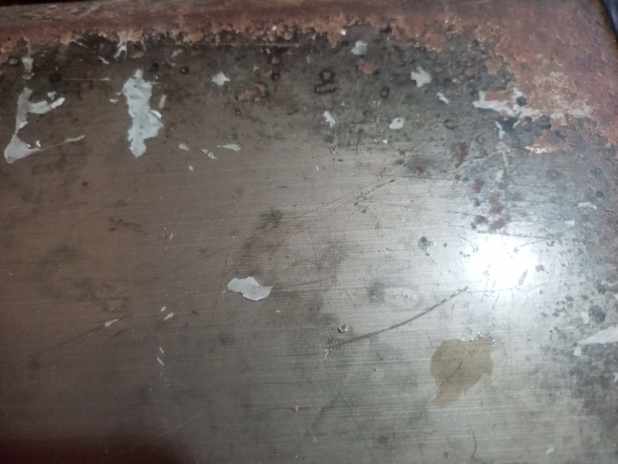 The image shows a close-up of a rusty and worn metal surface. The surface is a dull, metallic gray with significant areas of rust and corrosion, particularly along the top edge. There are also patches of what appear to be peeling paint or a coating, in irregular shapes and shades of white and light gray. Small dark spots, possibly dirt or more rust, are scattered across the surface. Some faint scratches and lines are visible, indicating wear and tear. A light brown stain is present in the lower right quadrant of the image. The overall impression is one of age and neglect. The location of this scene in Lhokseumawe, Indonesia, is not discernible from the image itself.