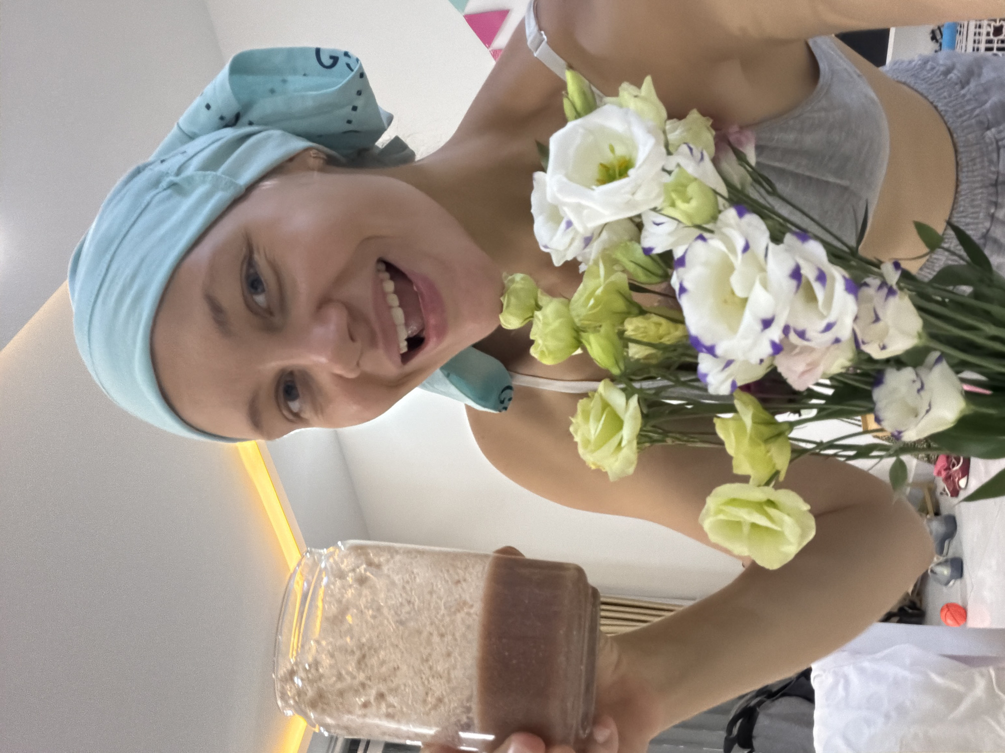 A young Caucasian woman wearing a light blue bandana and a grey sports bra is holding a glass jar filled with a brown beverage in her left hand and a bouquet of white and green flowers in her right hand. She is smiling broadly with her mouth open. The background appears to be an indoor setting with soft lighting, including an illuminated ceiling feature. The woman's clothing suggests a casual or relaxed environment. The image is framed from the chest up, with the focus on the woman and the objects she is holding. The location of Bac Nha Trang Ward, Vietnam, is not discernible from the visual information in the image itself.