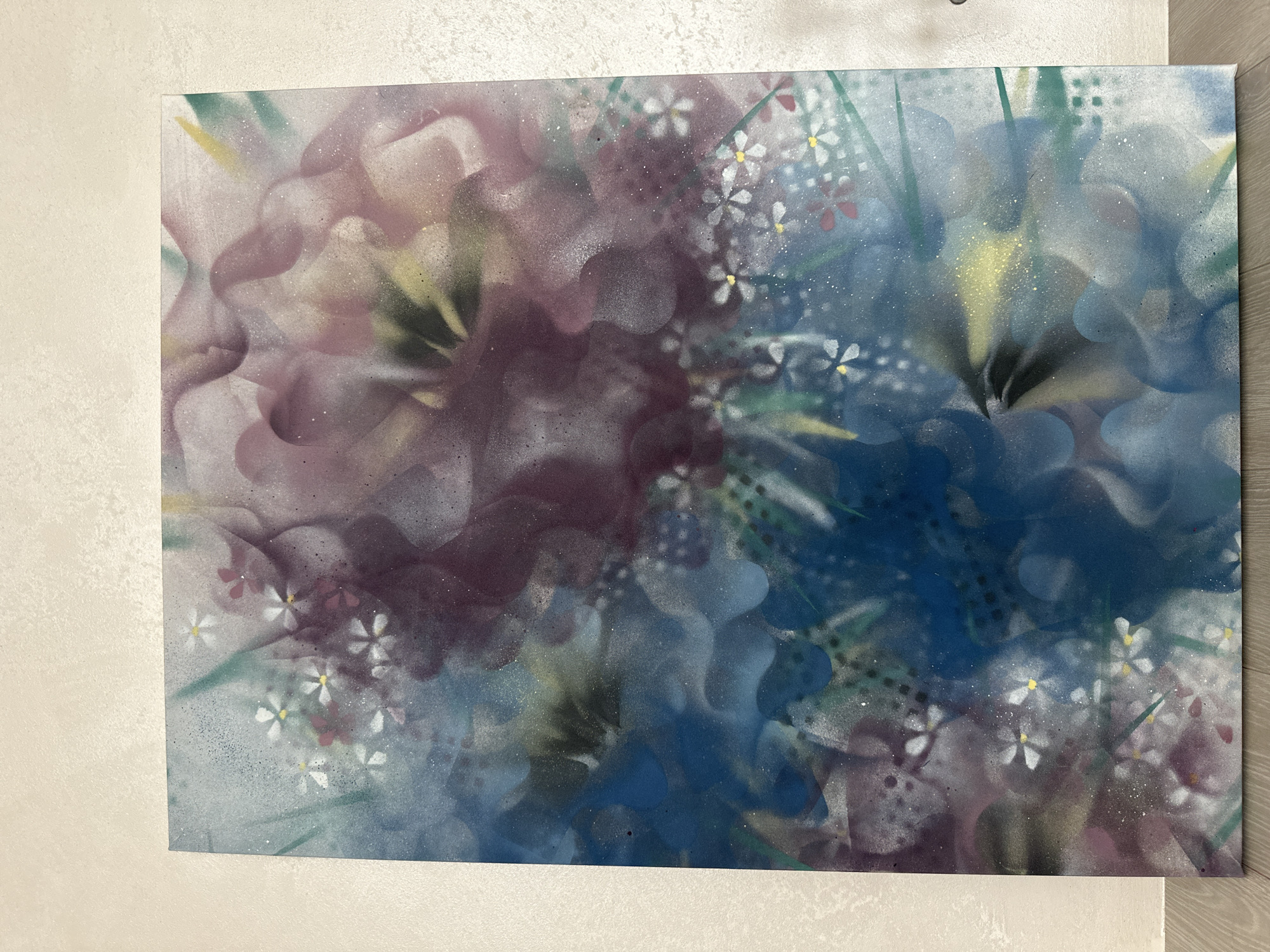 This is an image of an abstract floral spray-painted artwork leaning against a beige, textured wall. The painting is predominantly in shades of blue, purple, and white, with hints of yellow and green. Small white flowers with yellow centers are scattered throughout the composition. The artwork is displayed in what appears to be an indoor setting, possibly a residential or gallery space. No specific location in Minsk, Belarus can be identified from the visual information provided.