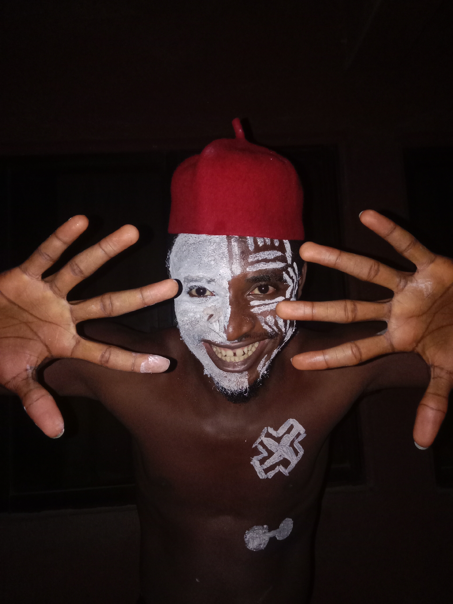 A medium shot captures a man with dark skin and a partially painted face, adorned in white paint. The right side of his face is covered entirely in white, while the left side features intricate white patterns. A red cap rests atop his head. His body is bare, except for white paint designs on his chest. He holds both hands up with fingers splayed, positioned on either side of his face. The background is dark and indistinct. The location is unknown.