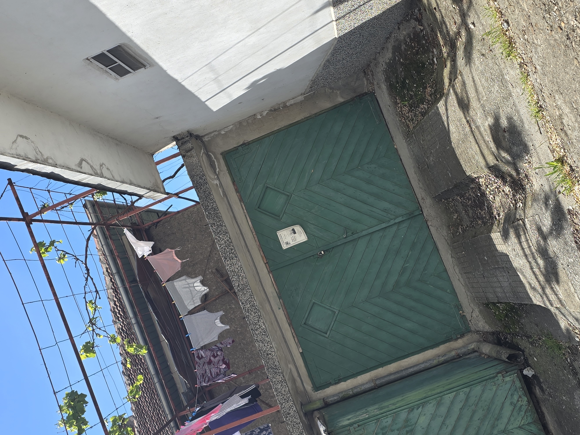The image shows an exterior shot of a building on a sunny day. The foreground is dominated by a two-door wooden garage painted green with a chevron pattern. A paper or flyer is affixed to the door. To the left, clothes are hanging on a line strung from a metal trellis-like structure covered with climbing vines against the building's exterior. The sky is visible in the background, suggesting an open space or yard. The building’s architecture has a utilitarian aesthetic. The location in the city is unknown.