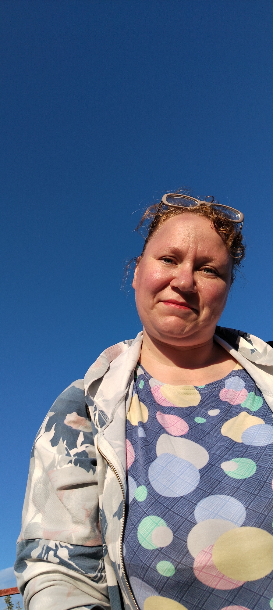 A woman is captured in a close-up, low-angle portrait, looking directly at the camera. She has reddish-blonde hair with glasses pushed up onto her head. Her face shows fair skin with rosy cheeks, and she wears red lipstick, exhibiting a subtle, closed-mouth smile.
She is dressed in a patterned t-shirt featuring a blue base with a grid texture and large, pastel-colored abstract shapes or circles in shades of pink, yellow, light blue, mint green, and white. Over this, she wears an unzipped jacket with an abstract floral or leaf pattern in muted tones of grey, white, and blue. The jacket's zipper is visible.
The background is a vast, clear, and bright blue sky, indicating an outdoor setting on a sunny day. Strong sunlight illuminates her, casting soft shadows under her chin and neck. The overall impression is one of casual contentment. There is no visible text in the image. The photo was taken in Chaikovsky, Russia.