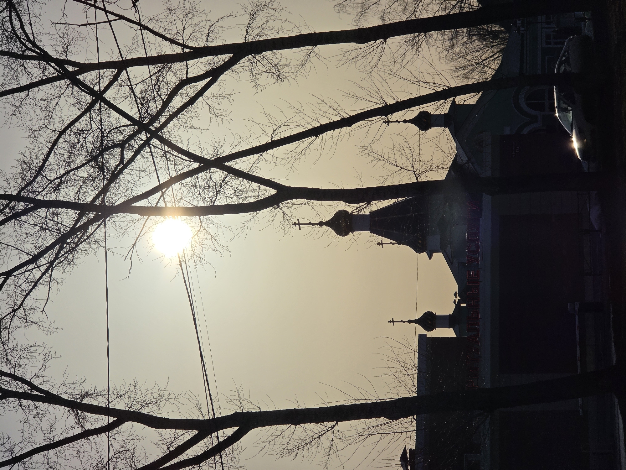 The image depicts a backlit urban scene in Chaikovsky, Russia, at either sunrise or sunset, characterized by a hazy, yellowish-white sky. The sun is prominently visible in the upper right, partially obscured by bare deciduous tree branches, causing a strong lens flare. Multiple leafless tree silhouettes dominate the foreground, with power lines crisscrossing between their branches.
In the midground, a Russian Orthodox church is visible, featuring a dark roof and at least three golden onion domes, each crowned with an Orthodox cross. Adjacent to the church, a commercial building displays illuminated red Cyrillic letters on its roofline, spelling "РИТУАЛЬНЫЕ УСЛУГИ" (Funeral Services). A partial sign on the church building identifies its location as "ул. Ленина 128" (Lenin Street 128). A light-colored sedan is parked in the lower right, its hood and windshield reflecting intense sunlight. The environment, marked by the bare trees, indicates a cold season. No individuals are discernible.