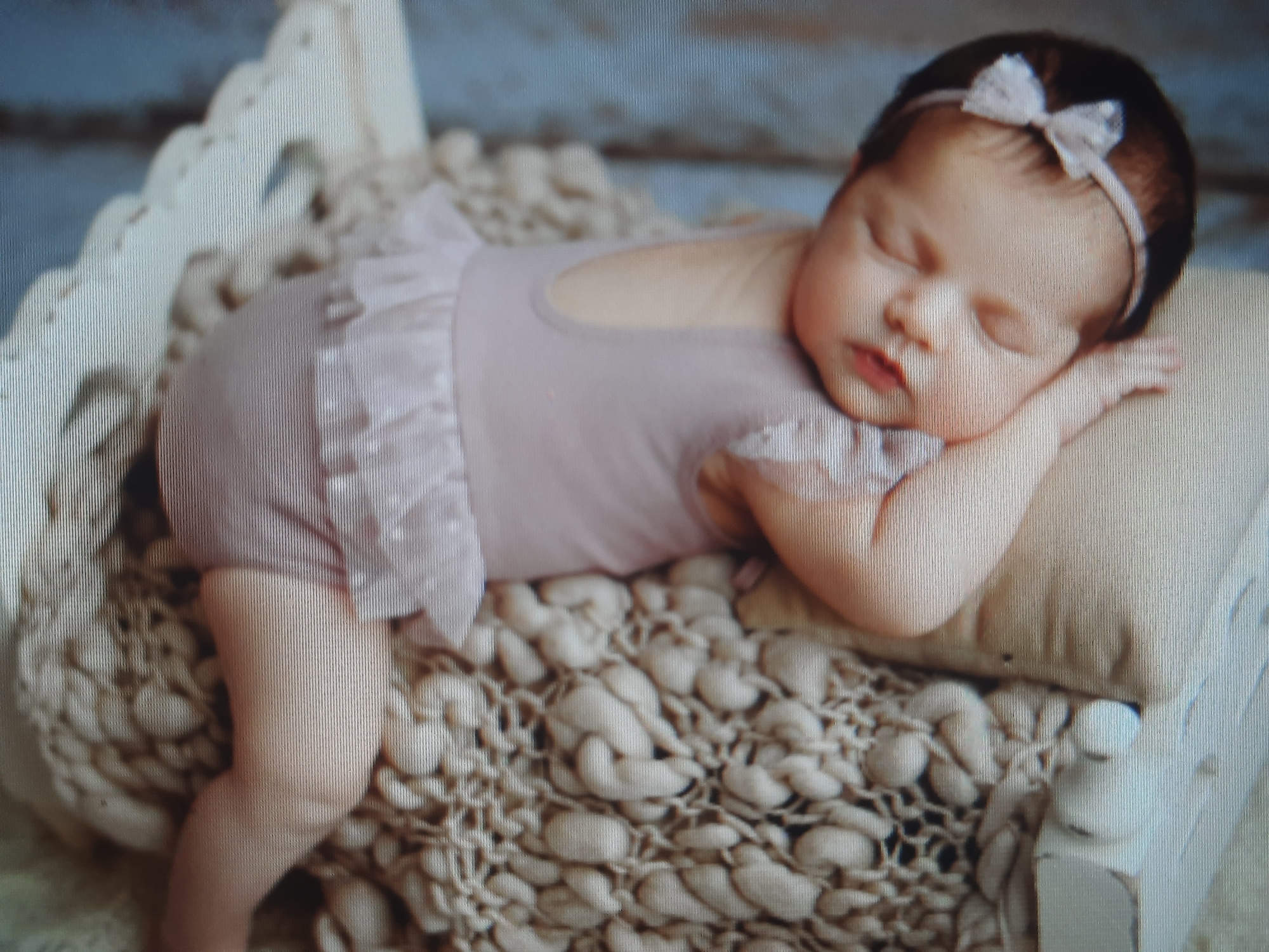 A newborn baby is lying on a textured, knitted surface, possibly a blanket or rug, within a decorative white cradle. The baby is wearing a light pink romper with ruffles and a matching bow in its hair. The baby's eyes are closed, and its head is resting on a beige cushion. The background is blurred, suggesting a studio or home environment. There is no indication of the city of Rosenheim, Germany.