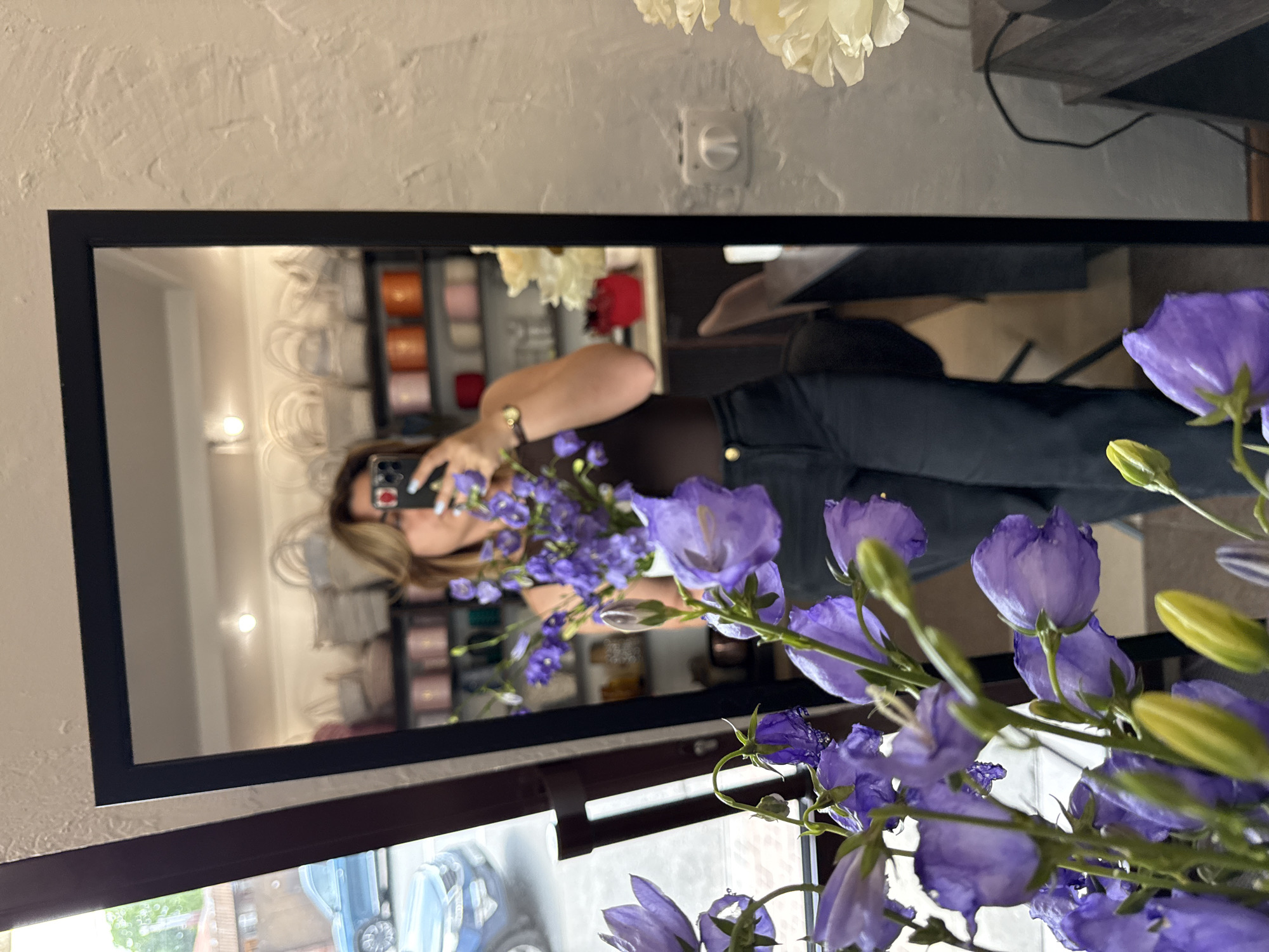 A woman is reflected in a tall mirror, holding a bouquet of purple flowers and a phone, taking a selfie. She is wearing a dark brown top and black pants. The mirror is framed by dark wood and is positioned in front of a textured light-colored wall. Behind her, shelves are visible in the reflection, stocked with various items, possibly in a shop. In the foreground, the bouquet of purple flowers is in focus. Outside the mirror, a blue car is visible through a window. The scene appears to be an indoor setting, possibly a flower shop or a room with retail displays.