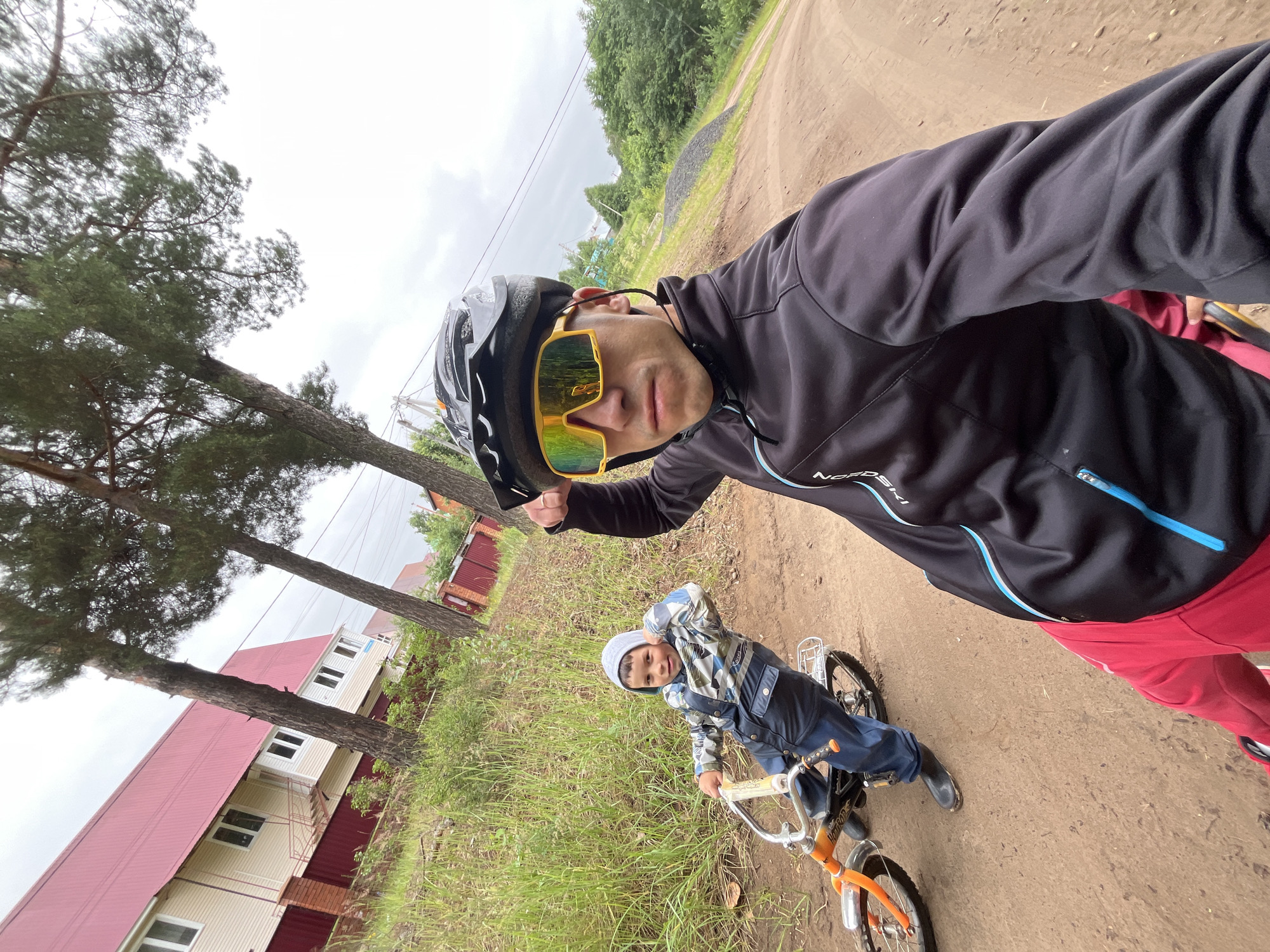 In Chaikovsky, Russia, on a dirt road bordered by lush green grass and trees, two individuals are engaged in outdoor activity. A man, wearing a black cycling jacket, red pants, a helmet, and yellow-tinted sunglasses, is captured in a selfie-style shot, with his right arm raised. To his left and slightly behind, a young boy on an orange and black bicycle, dressed in a patterned jacket and dark pants, is visible. The background features a house with a red roof and beige siding, a dark metal fence, and more trees under a cloudy sky. The overall scene depicts a casual recreational outing in a semi-rural or suburban setting.