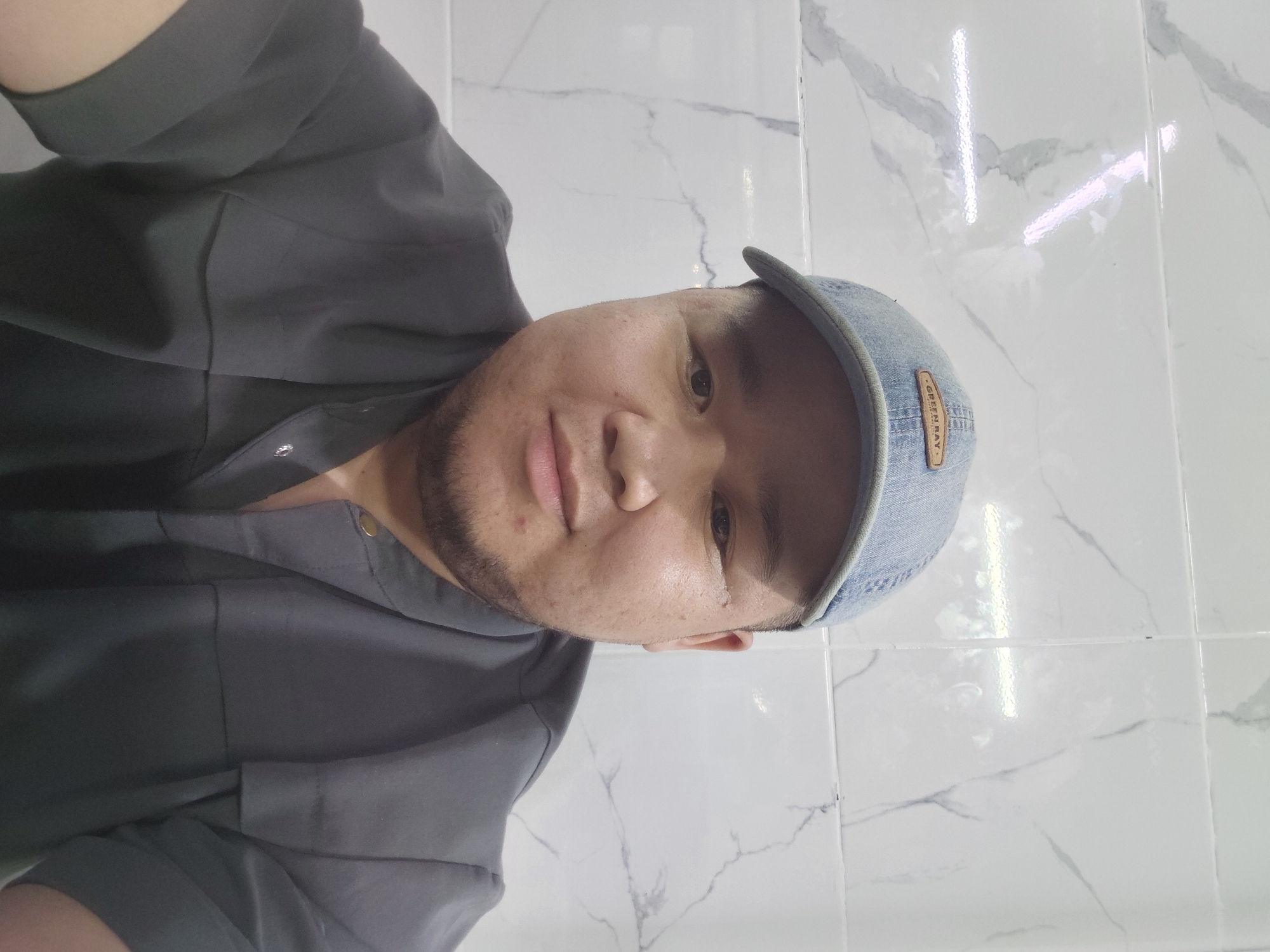 A portrait photograph shows a person wearing a light blue baseball cap with the text "GREEN BAY" visible on a patch. The individual is also wearing a dark grey collared shirt with short sleeves. The background consists of a wall tiled with a white marble pattern, with horizontal light fixtures visible reflecting on the surface. The image appears to be taken indoors, likely in a residential or commercial setting within the city of Aktau, Kazakhstan, though no specific landmarks or identifying features of the city are present in the scene.