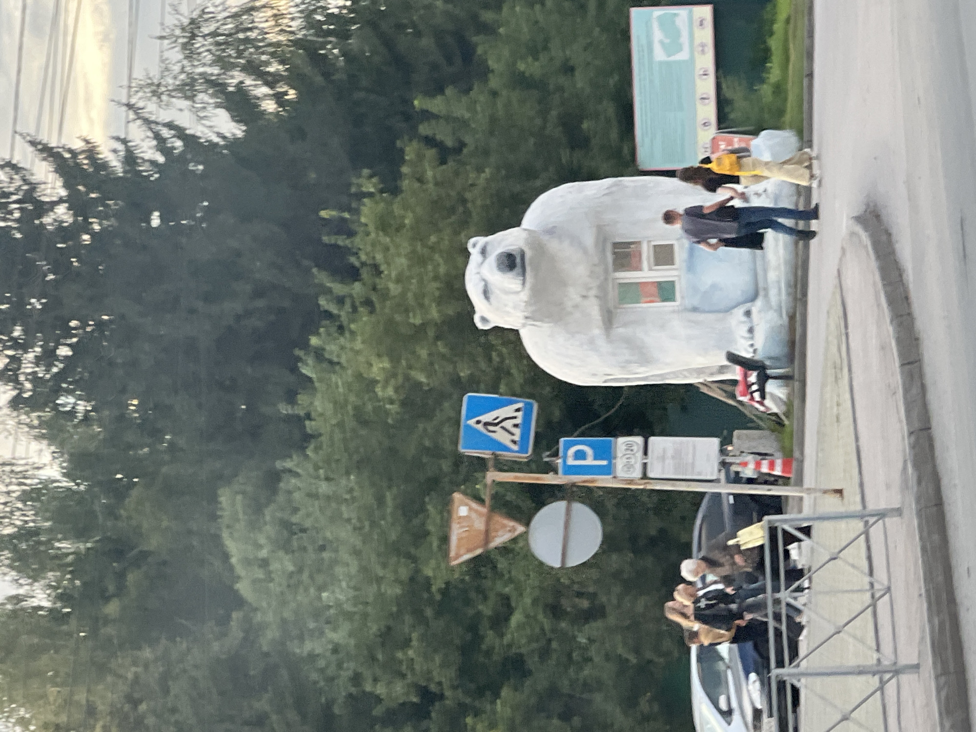 In Novosibirsk, Russia, a white polar bear statue with a window is situated near a road. Several people are visible in the vicinity. On the left, a group of individuals, including women and an older woman carrying a yellow bag, are walking near parked cars. Further to the right, a couple, a man and a woman with a yellow backpack, are walking away from the camera. Road signs, including a pedestrian crossing sign, a parking sign, and a warning sign, are posted near the statue. Trees and power lines form the background of the scene.